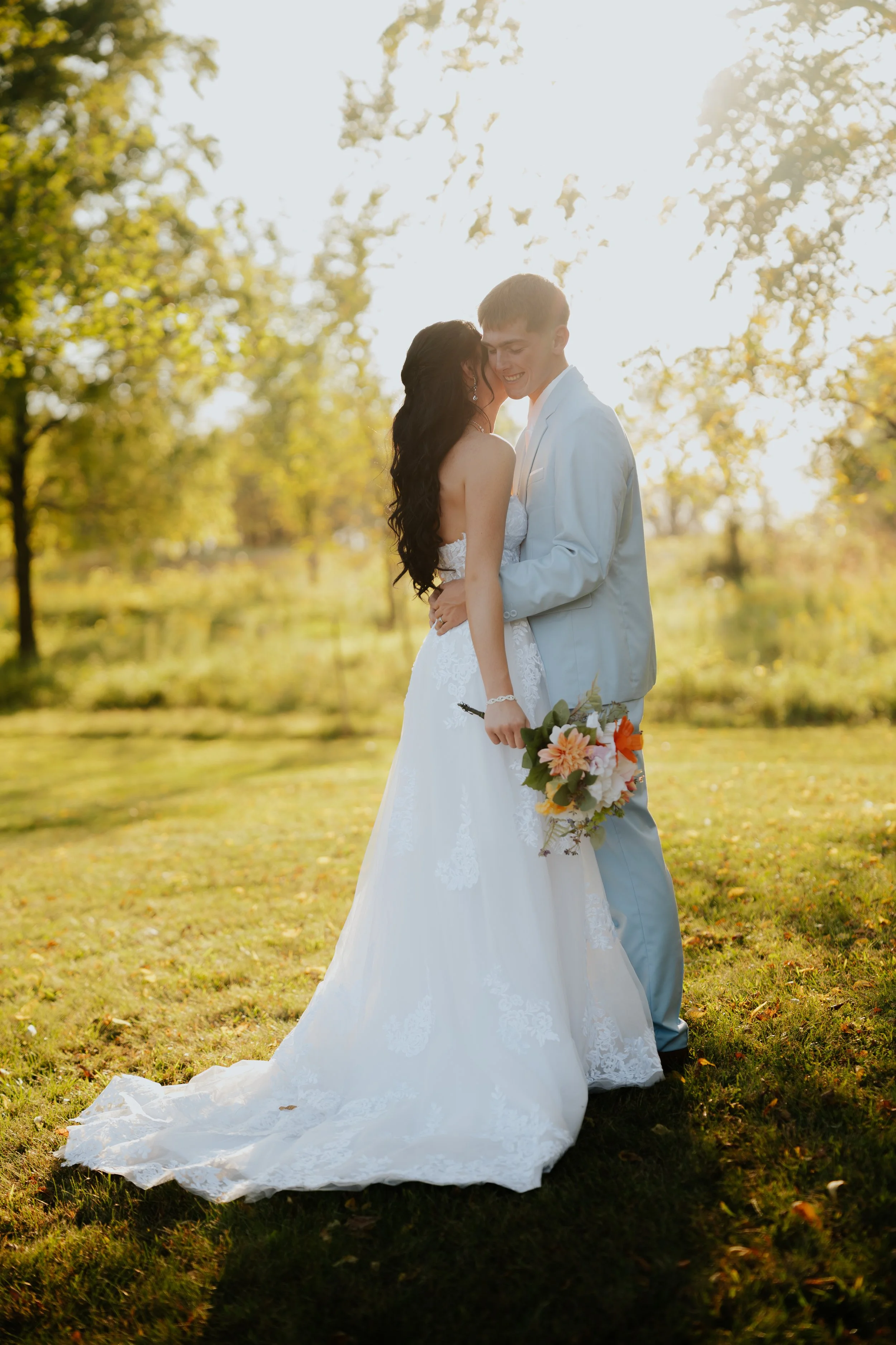 A newlywed couple shares a tender moment outdoors during sunset, with the bride in a white lace gown and the groom in a light gray suit, holding a bouquet of colorful flowers.