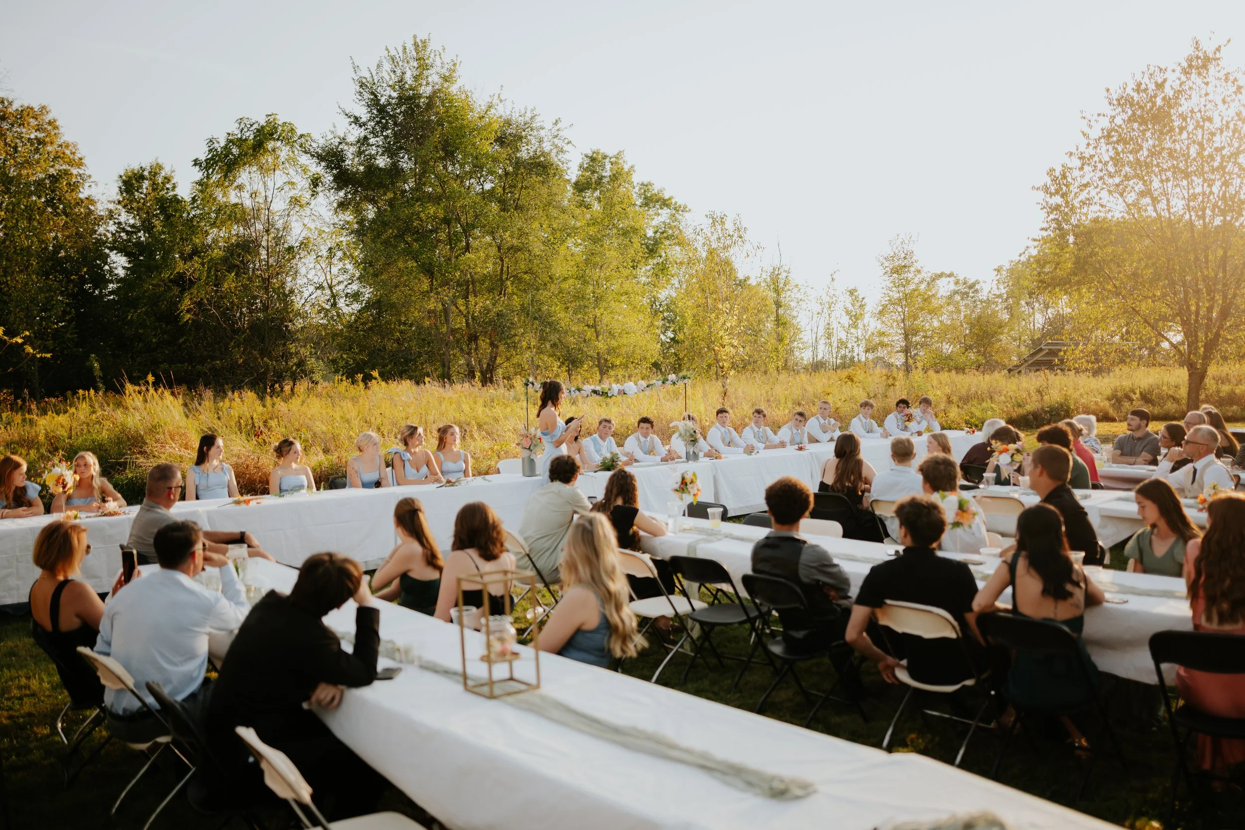 Outdoor wedding reception with guests seated at long tables, a woman giving a speech, and lush trees in the background during sunset.