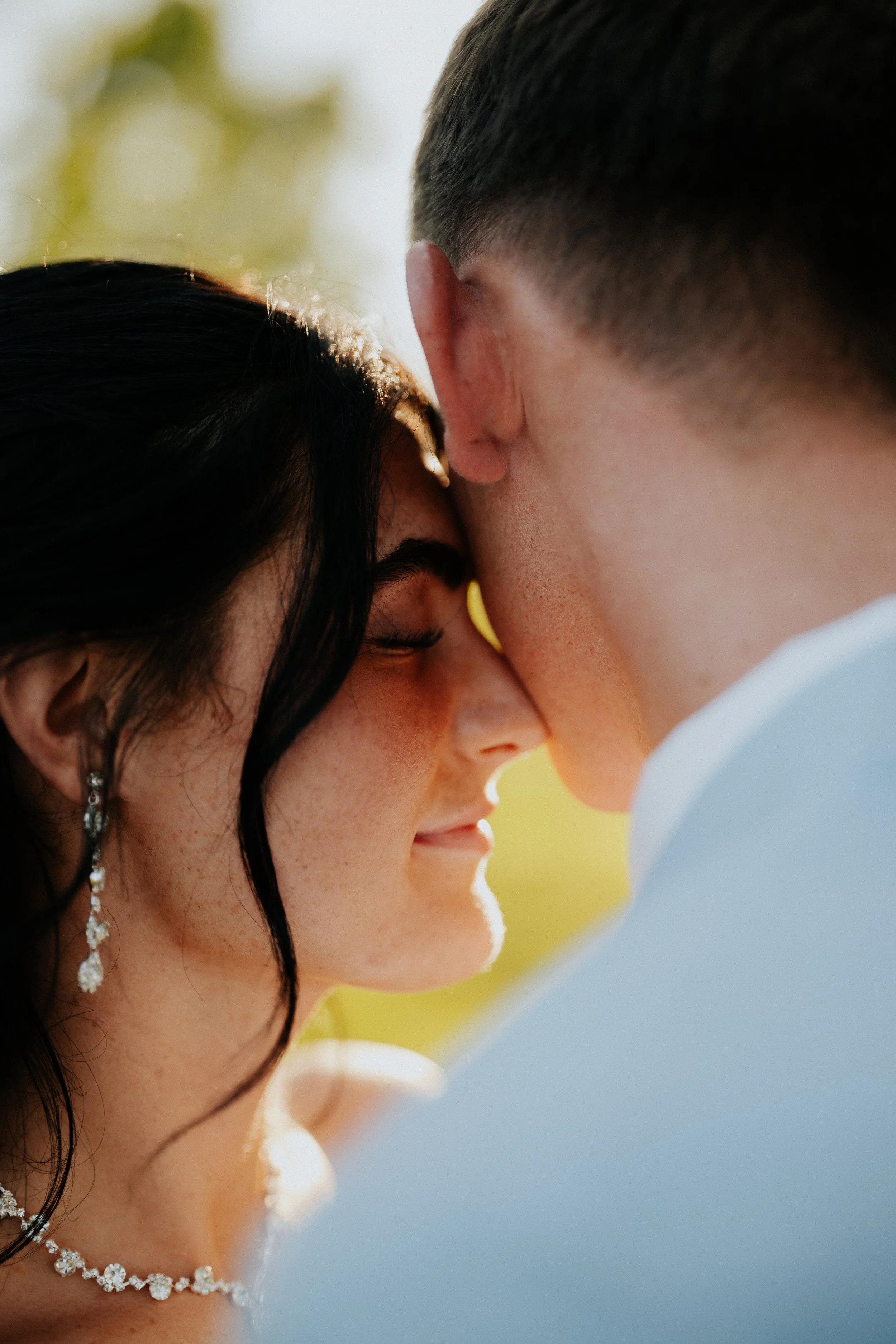 A couple is close together, with their foreheads touching, appearing to be sharing an intimate moment outdoors.