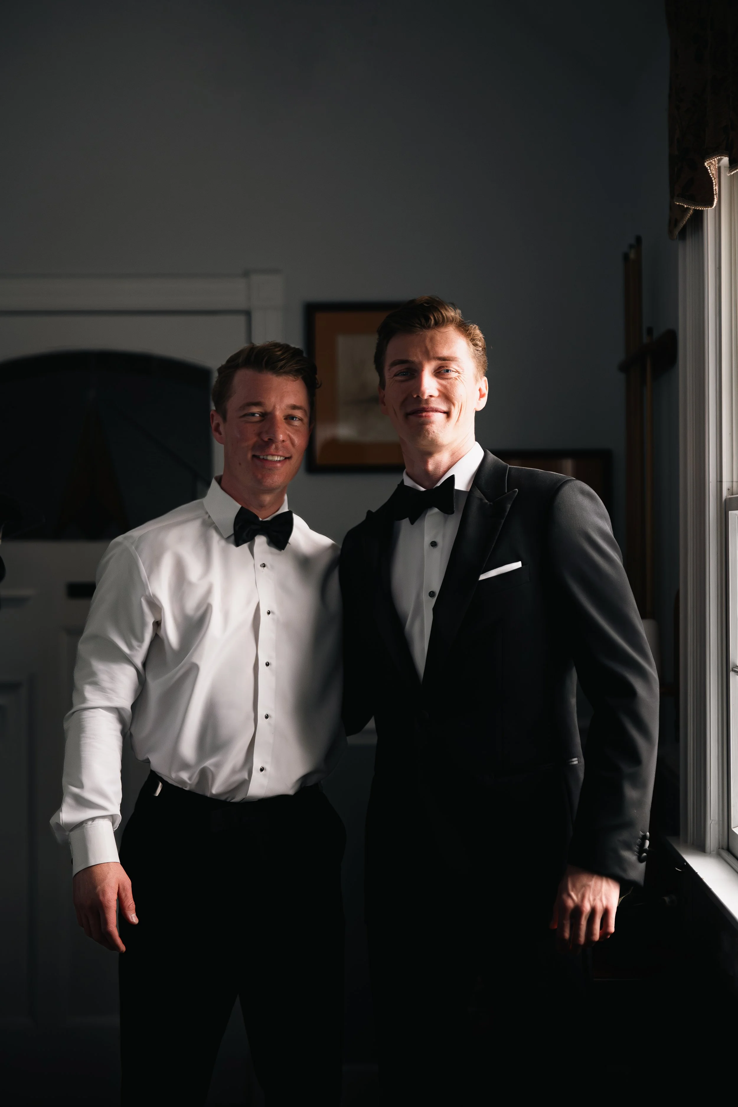 Two men in formal attire, one in a white shirt and bow tie, and the other in a black tuxedo, standing together indoors.