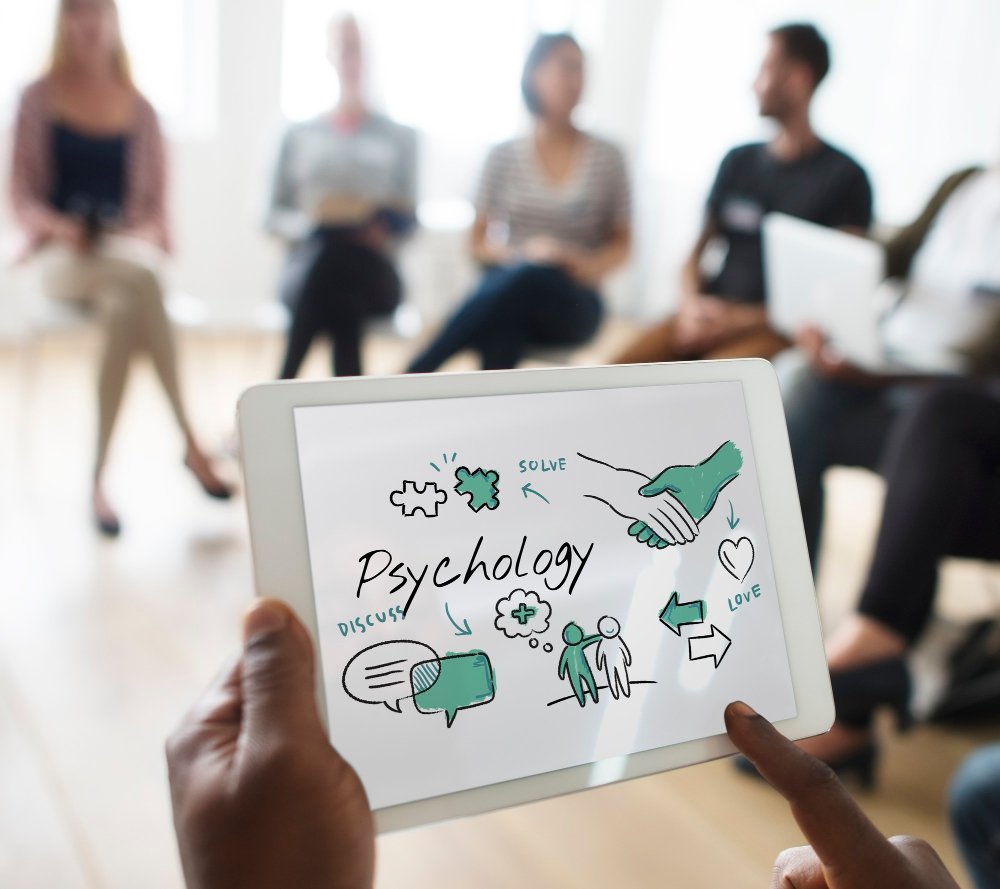Person holding a tablet displaying a psychology-themed illustration with words like "solve," "discuss," and "love." Blurred background of people sitting in a circle in a therapy or discussion setting.
