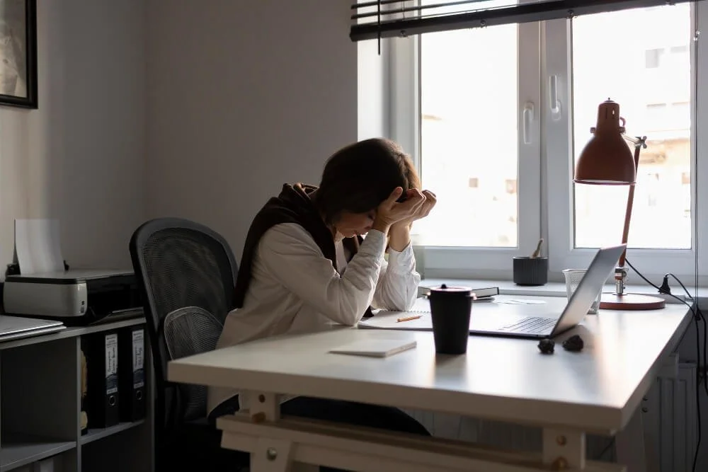 Person sitting at a desk, looking stressed, with hands on forehead in a home office setting. Laptop, coffee cup, and documents are on the desk. Sunlight is coming through the window.