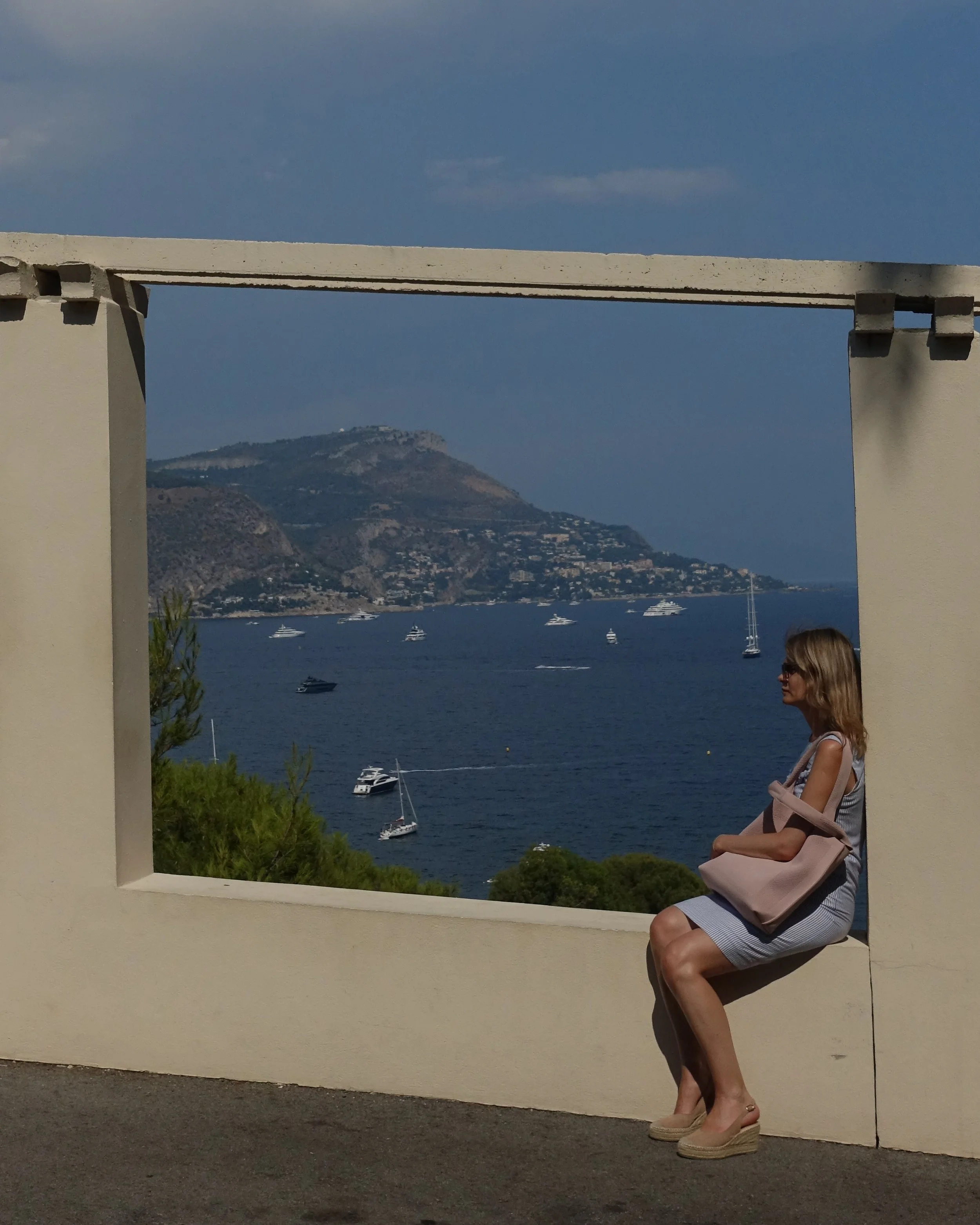 Une femme assise sur une rambarde en pierre, regardant vers la mer avec des bateaux naviguant devant des collines montagneuses, lors d'une journée ensoleillée.