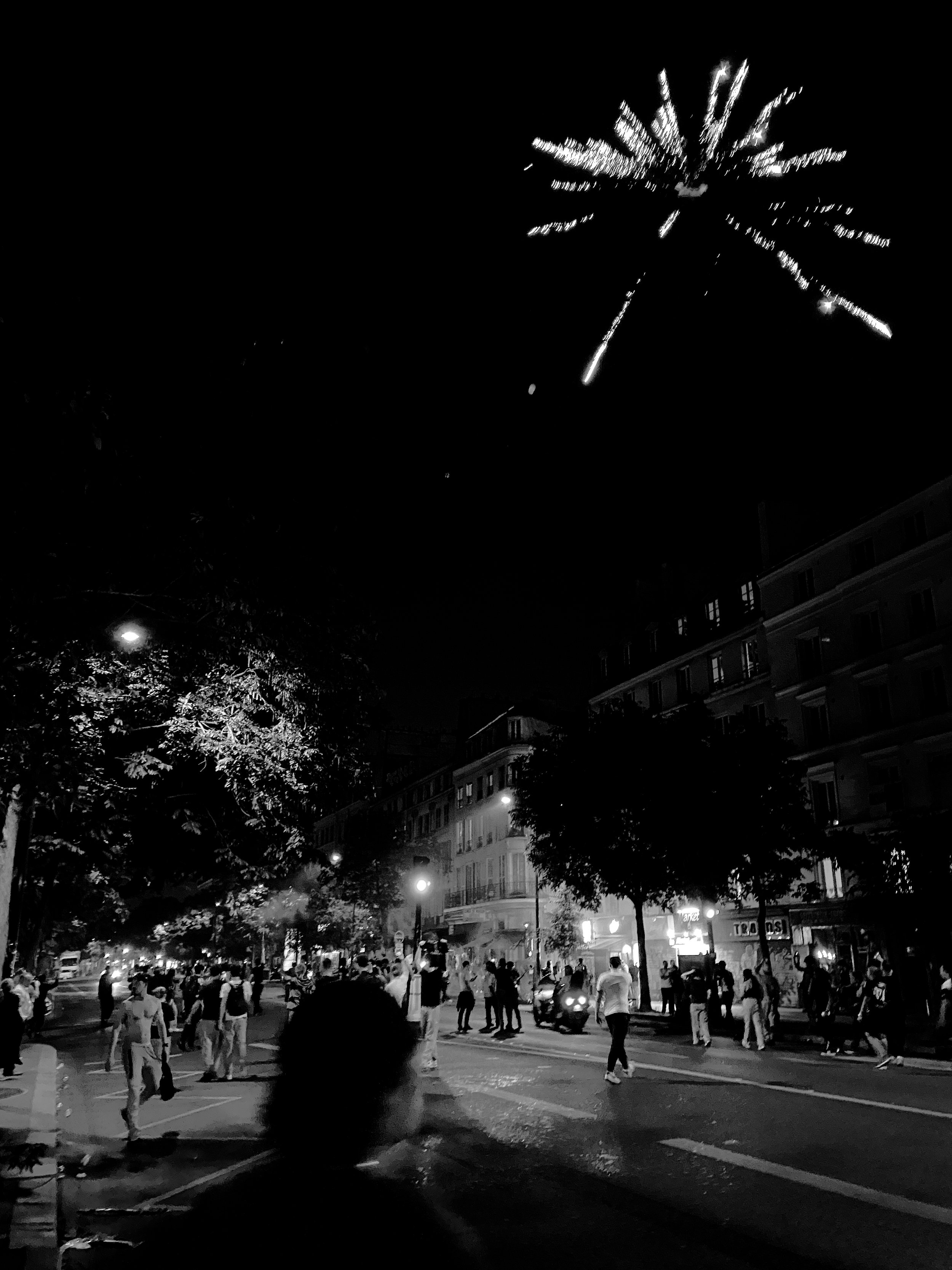 Feu d'artifice nocturne au-dessus d'une ville animée, avec des gens qui marchent dans la rue, des arbres et des bâtiments.