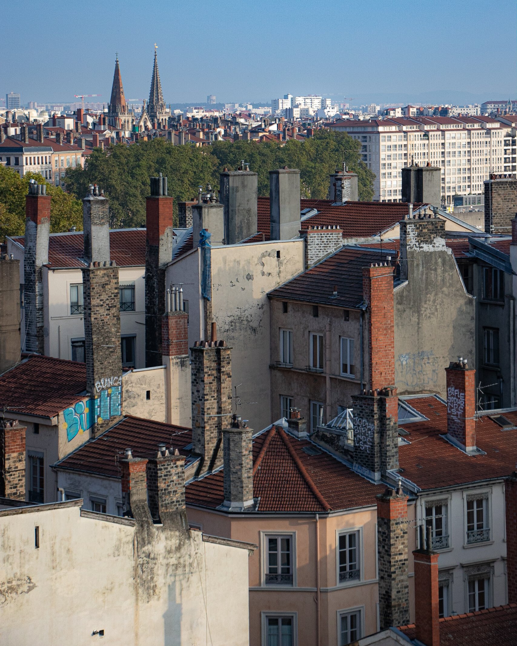 Toit de bâtiments parisiens avec cheminées en brique, graffiti sur un mur et tours de cathédrale en arrière-plan.