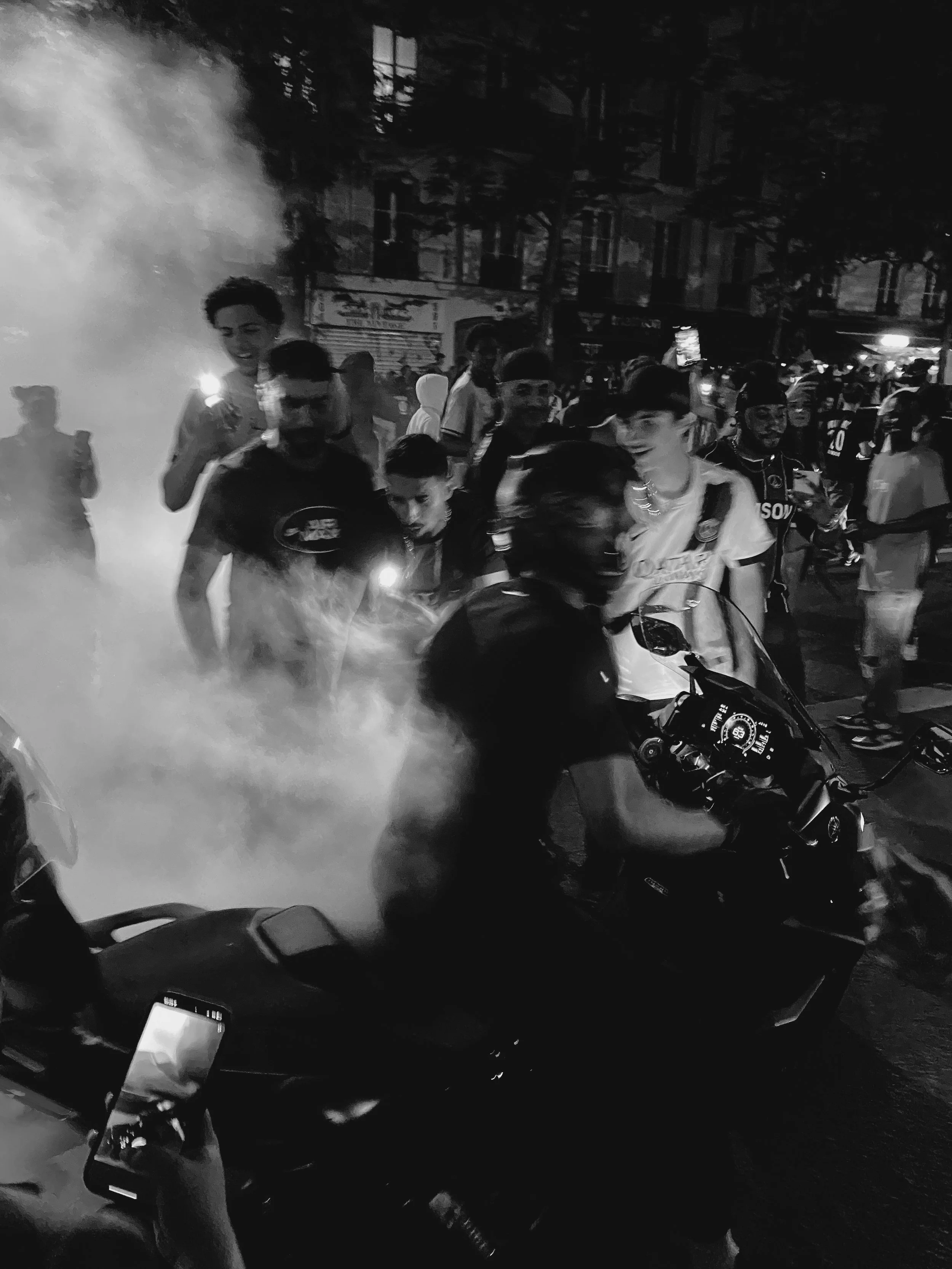 Une foule de jeunes gens dans une rue, certains portant des maillots de football, d'autres prennent des selfies ou regardent, avec de la fumée ou de la vapeur blanche dans l'air.