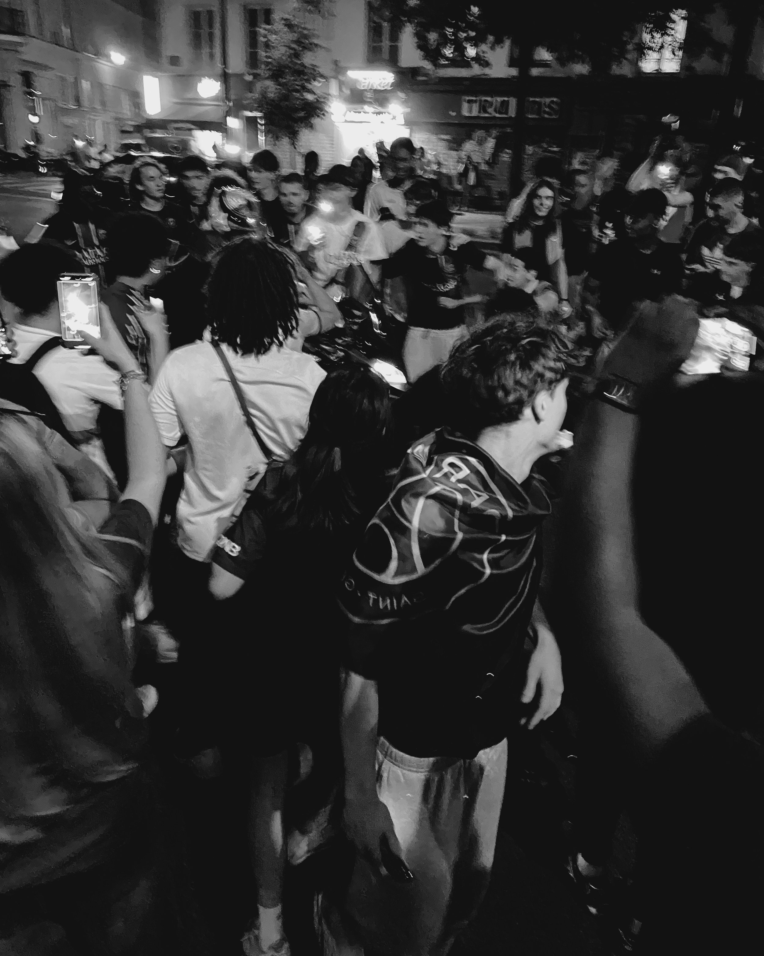 Une grande foule de personnes réunies dans une rue la nuit, certaines prennent des photos ou des vidéos avec leurs téléphones, ambiance de rassemblement ou d'événement public.