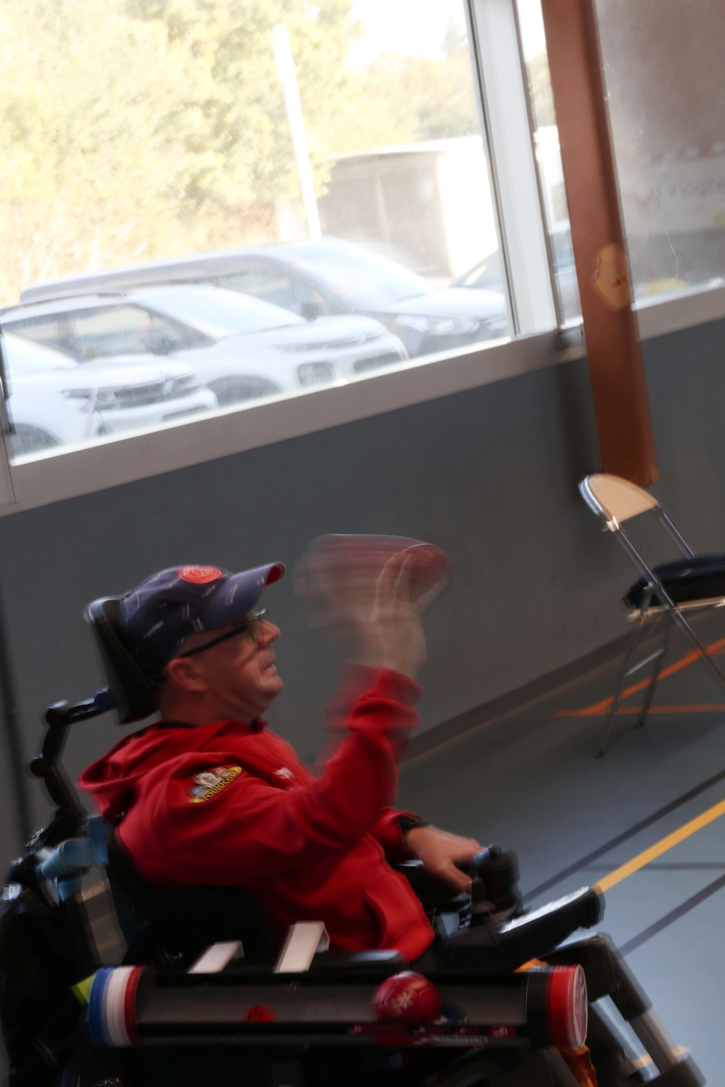 Un homme en fauteuil roulant, portant une casquette et une veste rouge, semblant faire un geste de la main à l'intérieur d'un bâtiment près d'une fenêtre avec des voitures garées dehors.
