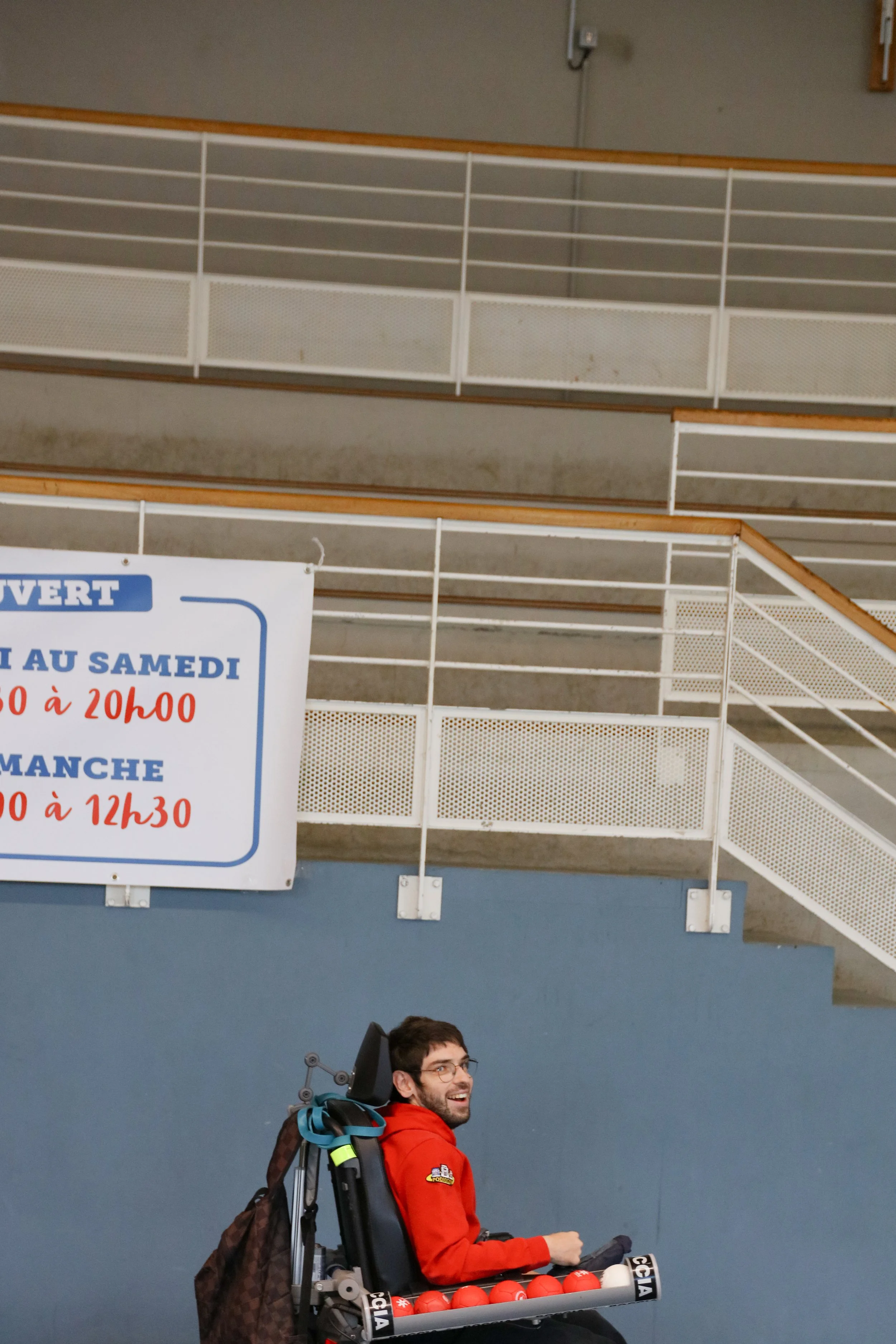 Un homme en fauteuil roulant dans une salle, portant un sweat rouge, souriant, avec une unité de contrôle pour cécité et une rangée de balles de tennis, dans une salle sportive.