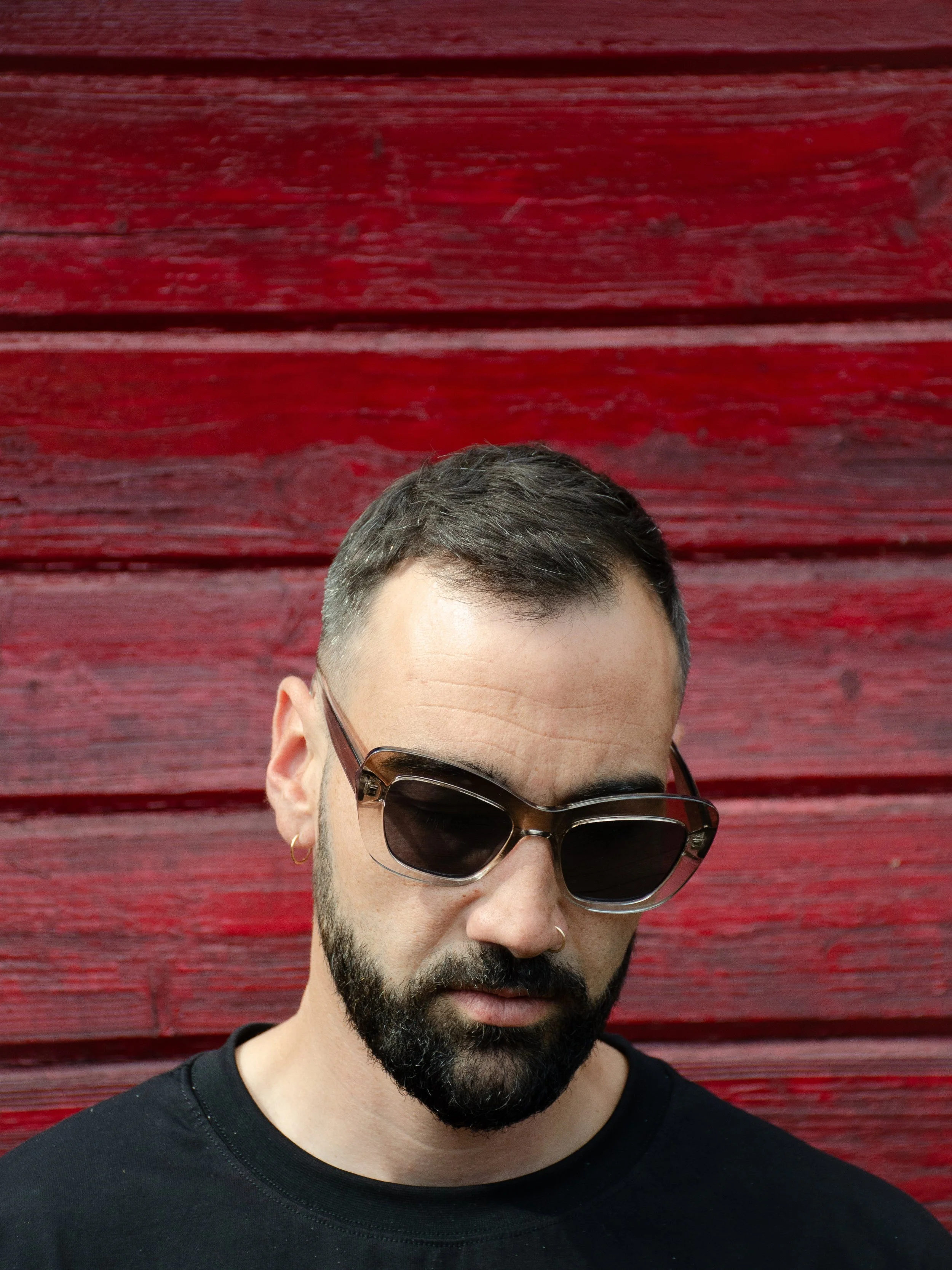 Homme avec barbe, portant des lunettes de soleil, écoutant attentivement contre un mur en bois rouge.