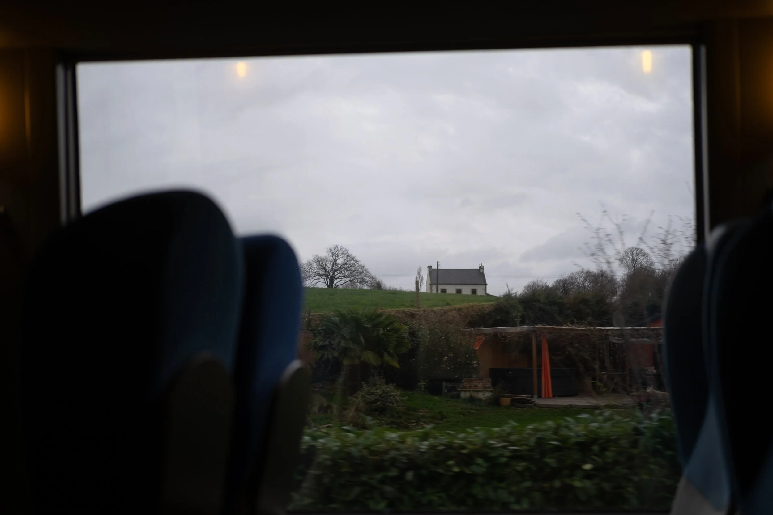 Vue depuis un autobus montrant une maison blanche au sommet d'une colline, entourée d'arbres et de végétation, avec un ciel nuageux.