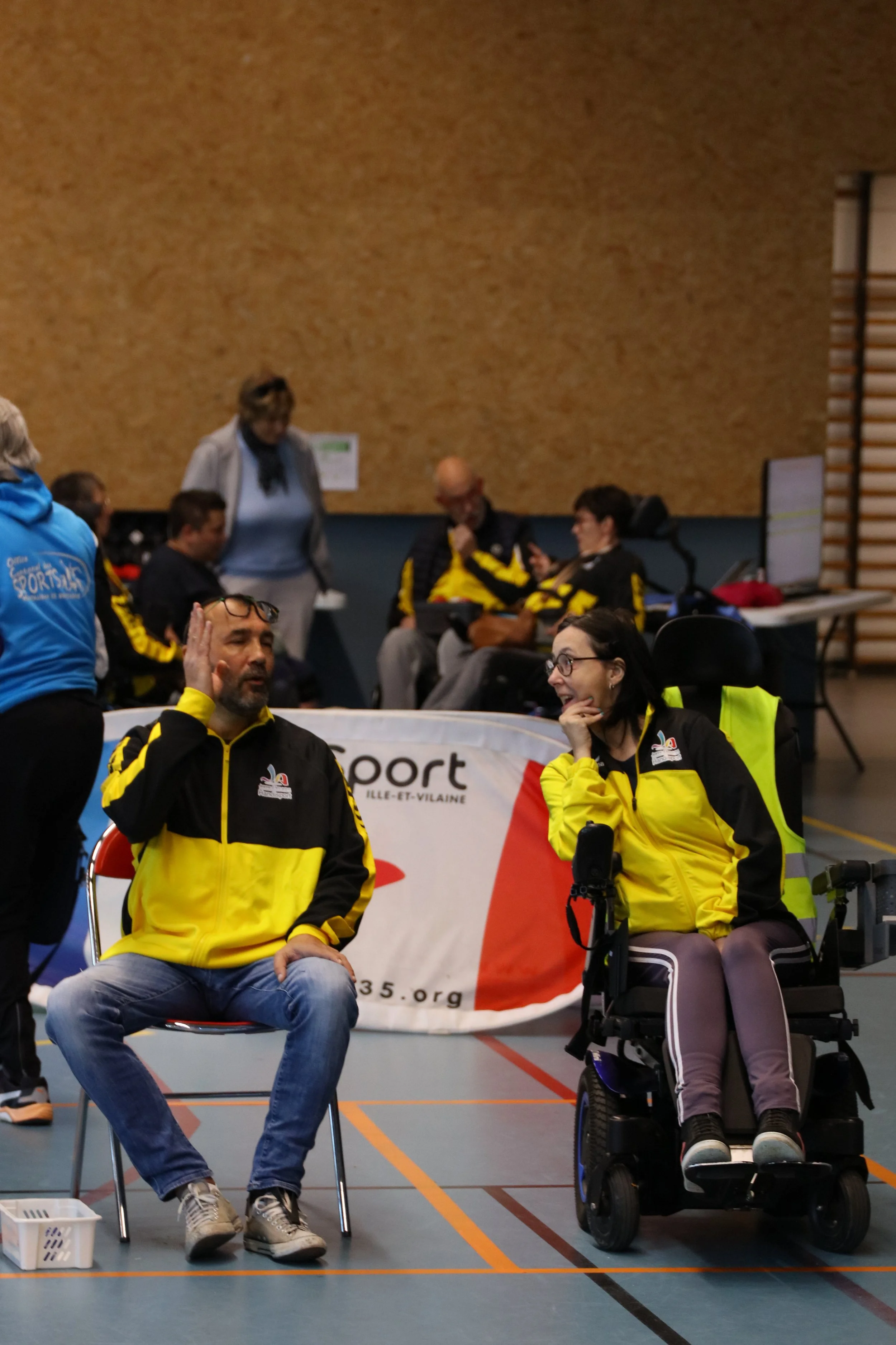 Une réunion ou un événement sportif avec des personnes portant des vestes jaunes et noires, certains assis et d'autres discutant, dans une salle intérieure.