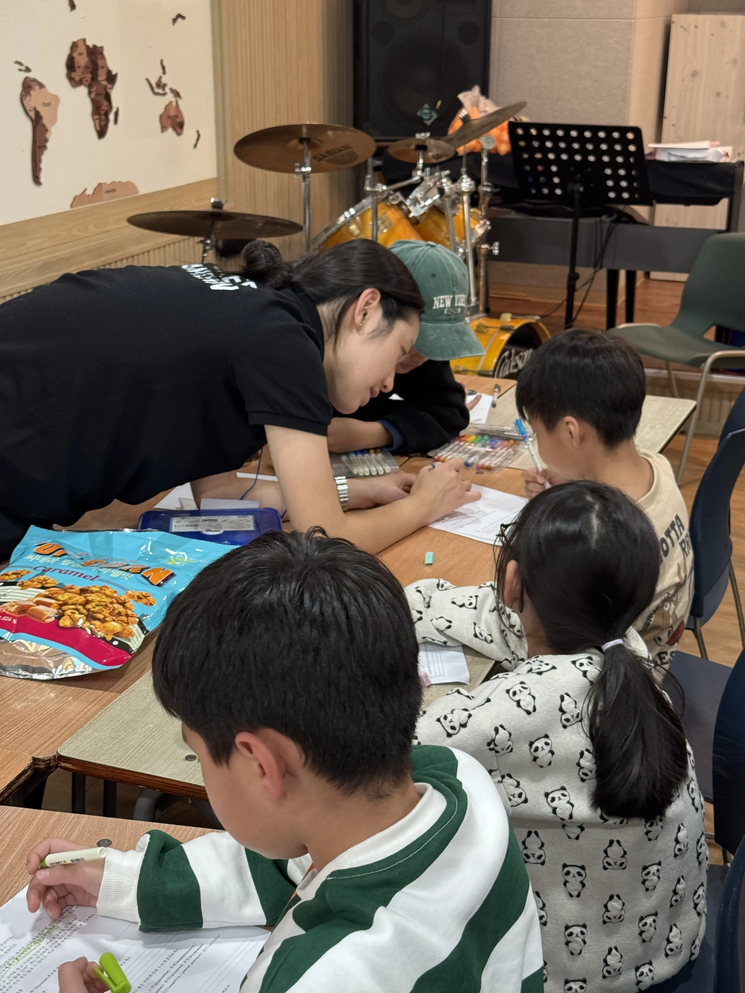 A group of children and a young woman engaged in a learning activity at a wooden table, with snacks and colorful pens, in a room with musical instruments and a world map on the wall.