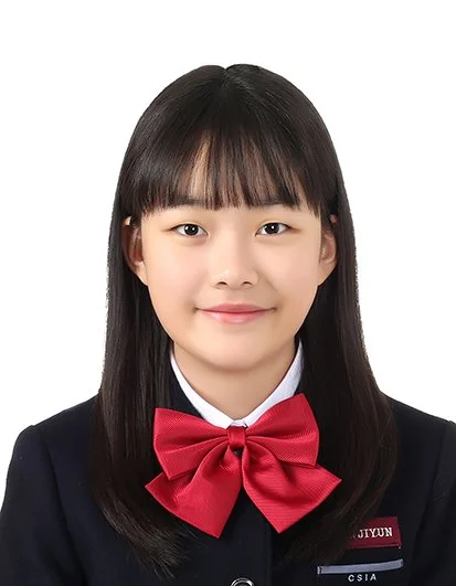 A young girl with long black hair, wearing a black school uniform and a large red bow tie, smiling against a white background.