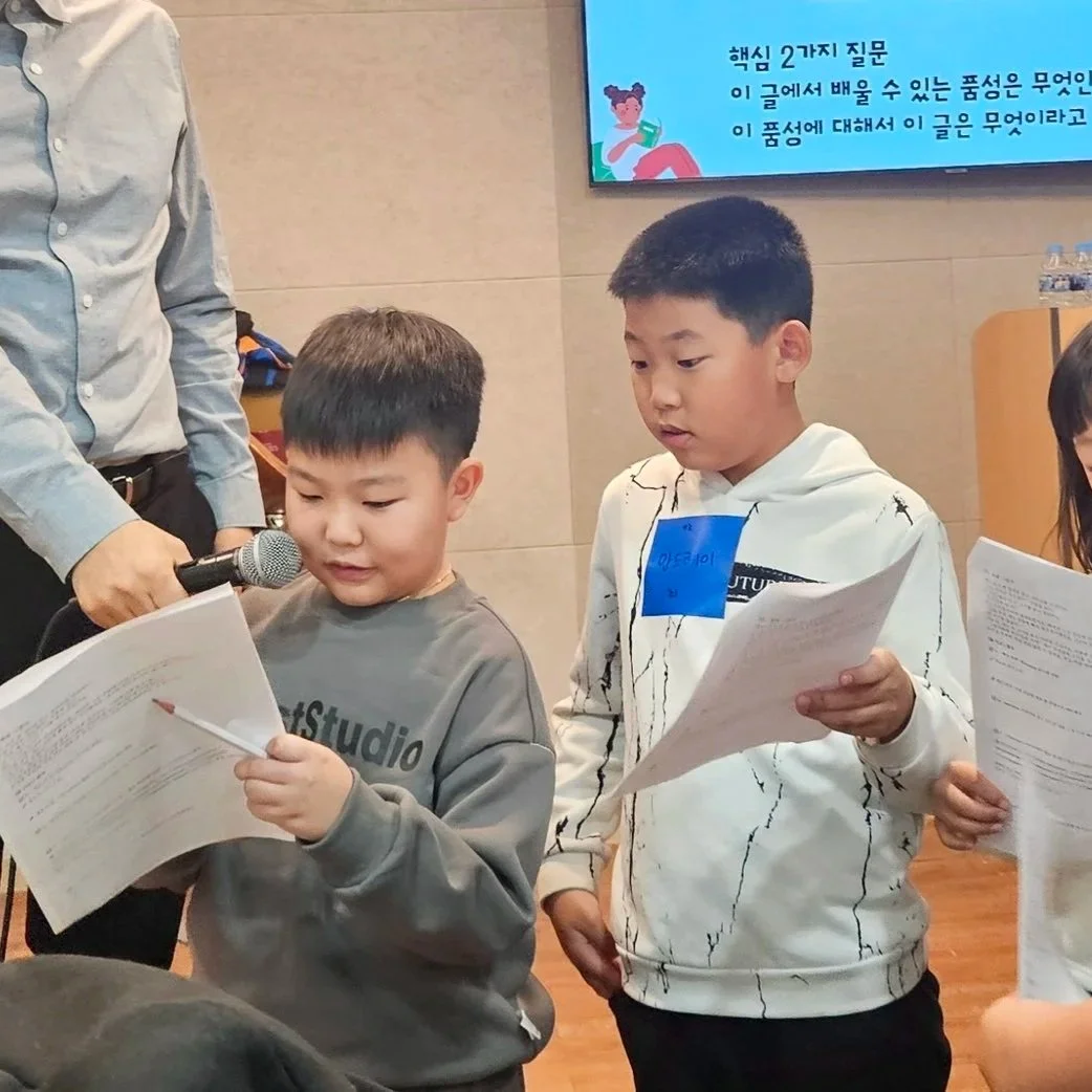 Two boys are standing in a classroom, reading from papers. A man is holding a microphone towards the boy wearing a gray sweatshirt. In the background, a screen displays text in Korean.