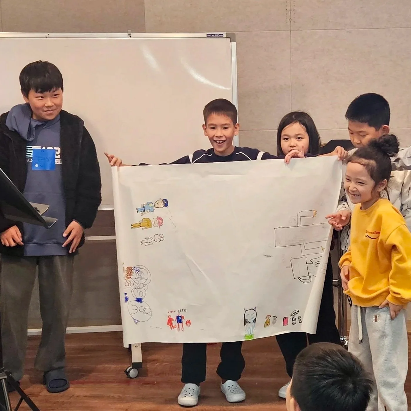 Group of children smiling and holding a large piece of paper with drawings and doodles, standing in front of a whiteboard.
