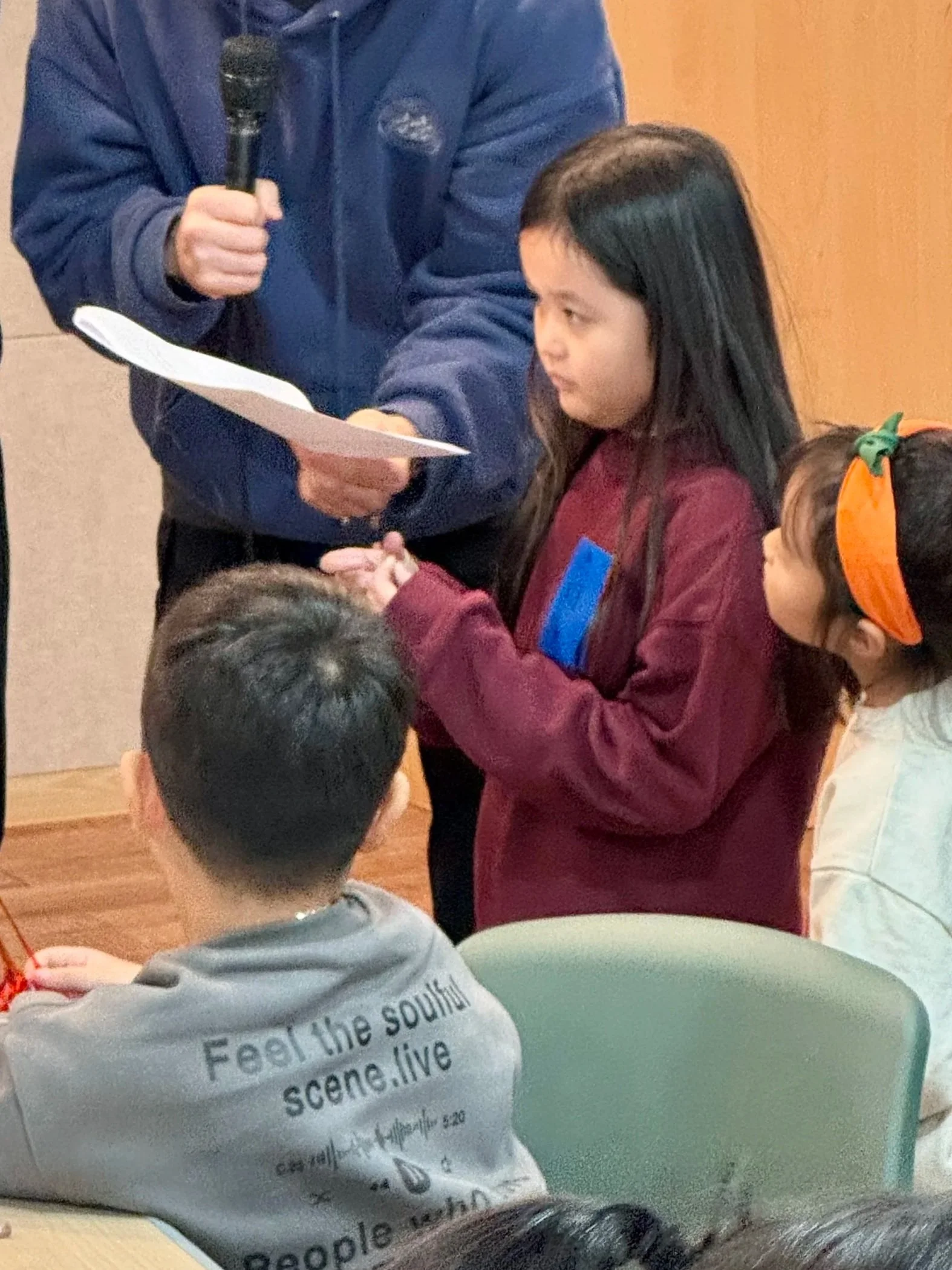 A young girl in a maroon sweatshirt is standing and holding a piece of paper, speaking to children seated at a table, with a person holding a microphone nearby.