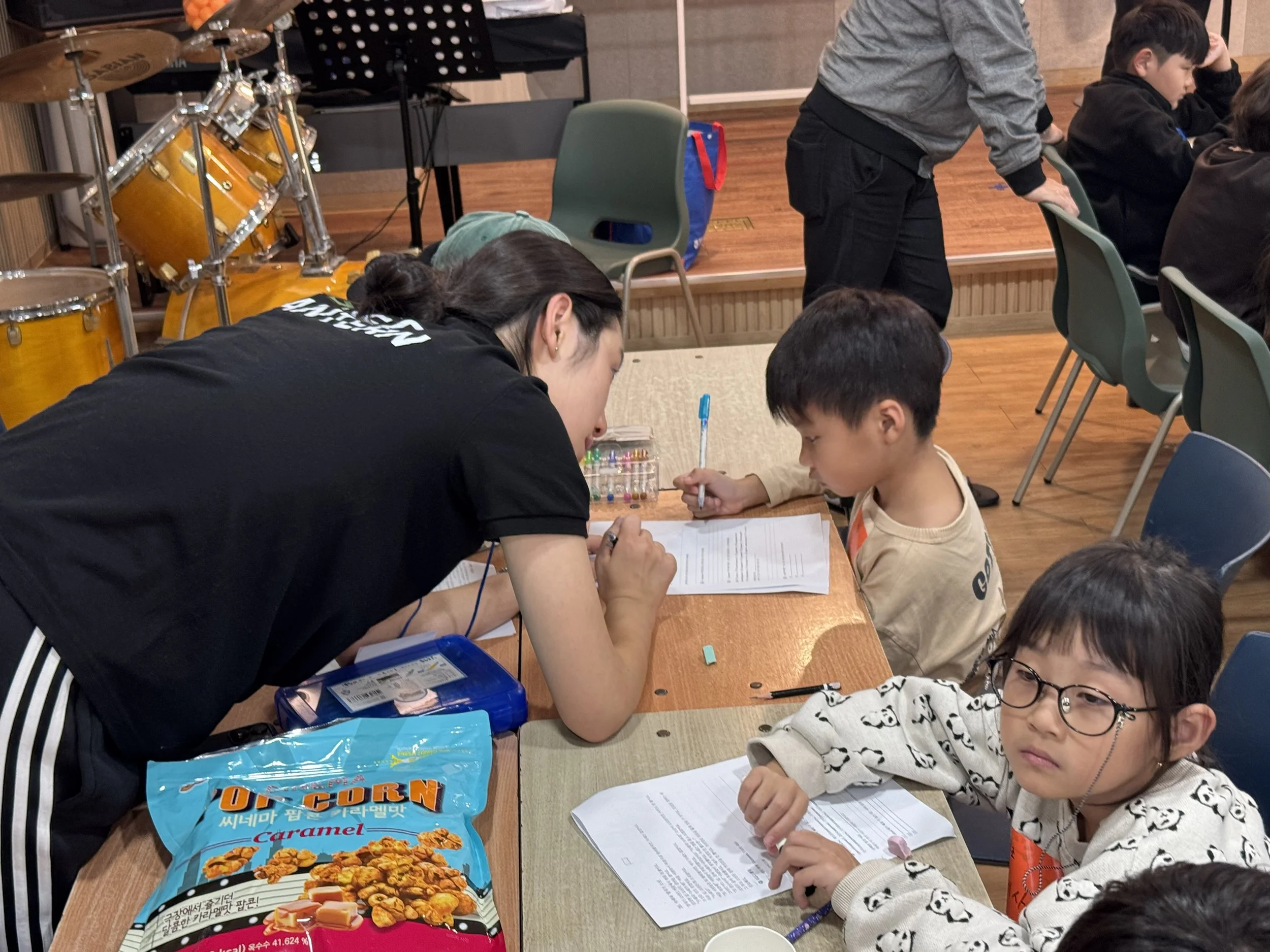 A young woman leaning over a table, assisting children with their work. Two children are visible, one of whom is writing in a notebook and the other looking at the camera with glasses. There is a bag of caramel popcorn and school supplies on the table, with more children and adults in the background in a room with wooden floors and chairs.