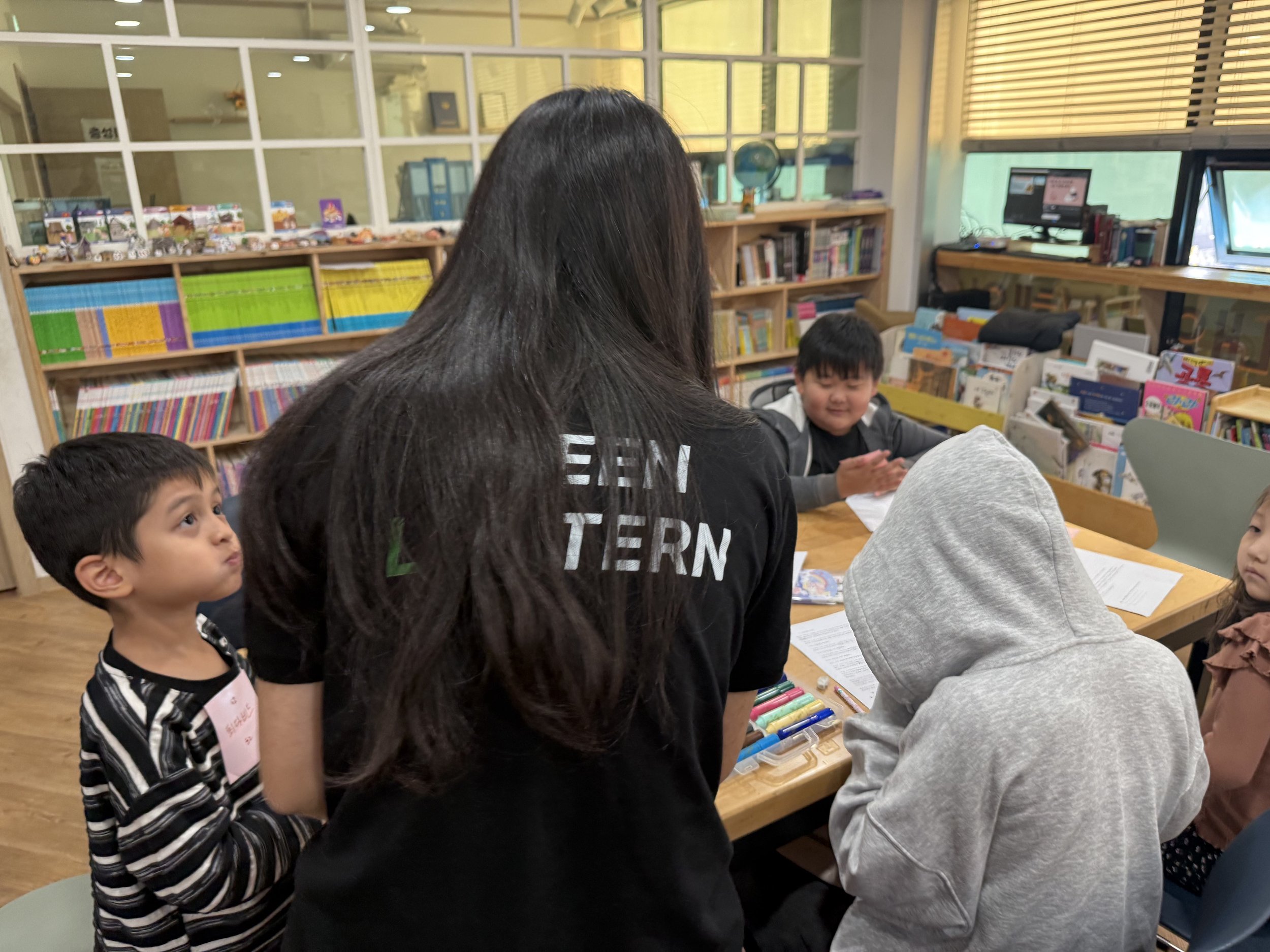 A woman with long black hair, wearing a black t-shirt, is standing at a table with four children in a classroom or library setting. The children are sitting around the table with books and colored markers. The background includes bookshelves filled with colorful books, educational supplies, and windows with blinds.