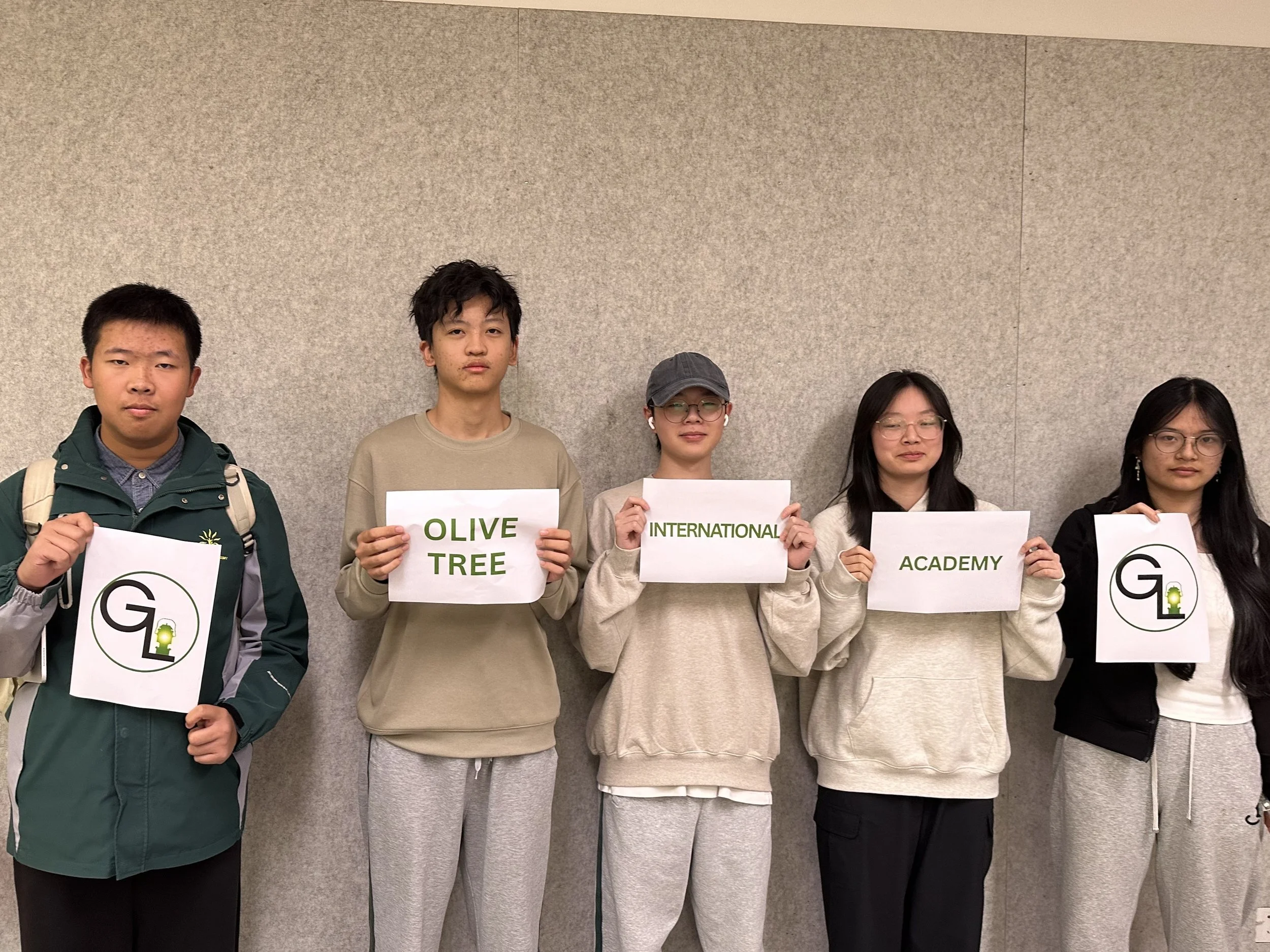 Group of five young people standing against a beige wall, holding signs that spell out 'G OLIVE TREE INTERNATIONAL ACADEMY' with some signs featuring the academy's logo.