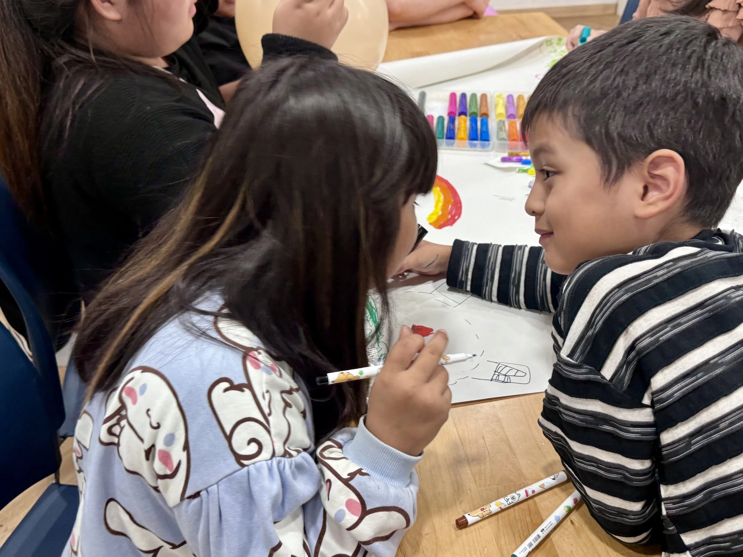 Children gathered around a table drawing with colored markers, with some drawings and markers visible on the table.