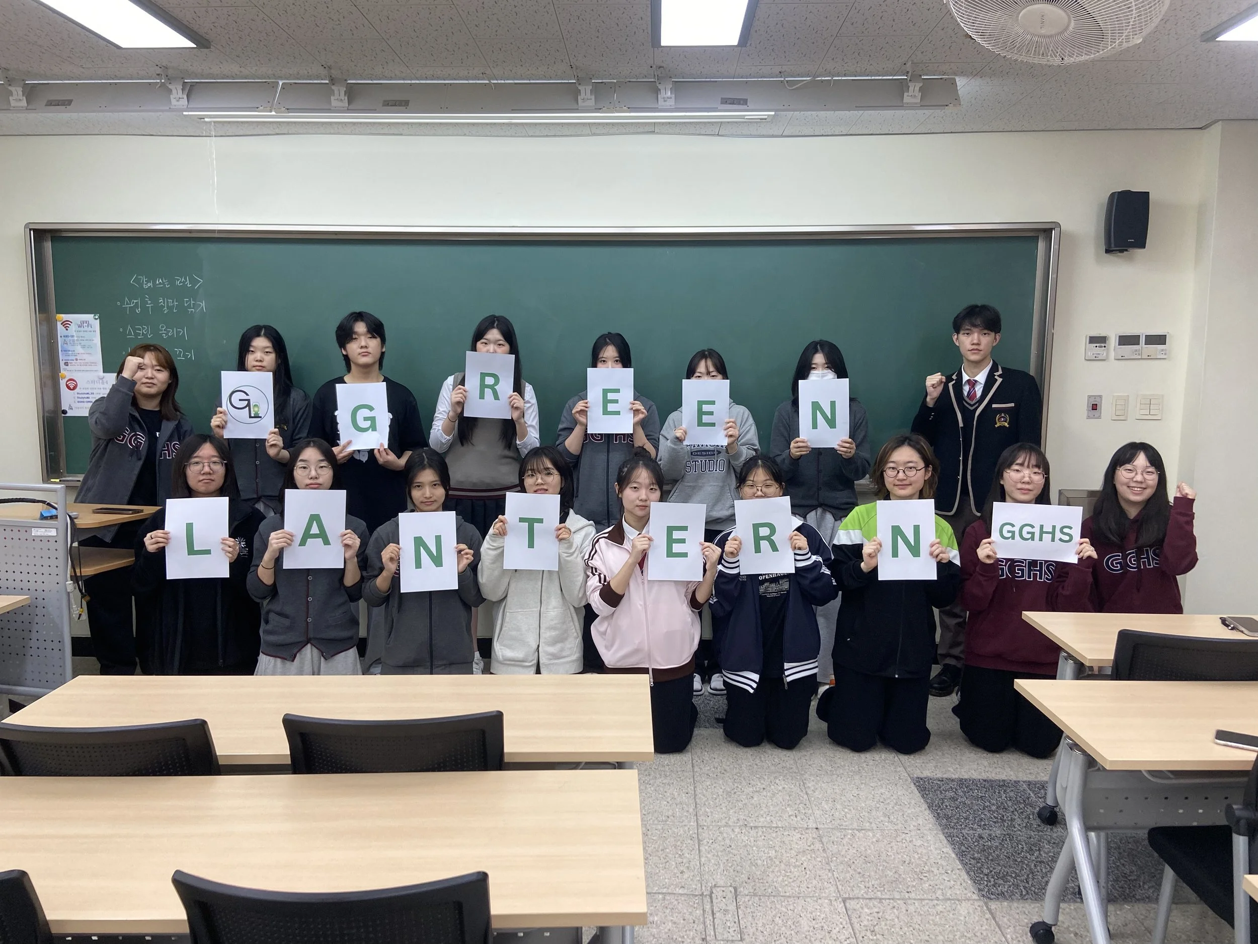 Group of students in a classroom holding signs that spell out 'GREEN LANTERN' and 'GGHS' with some students kneeling in front and others standing behind a chalkboard.