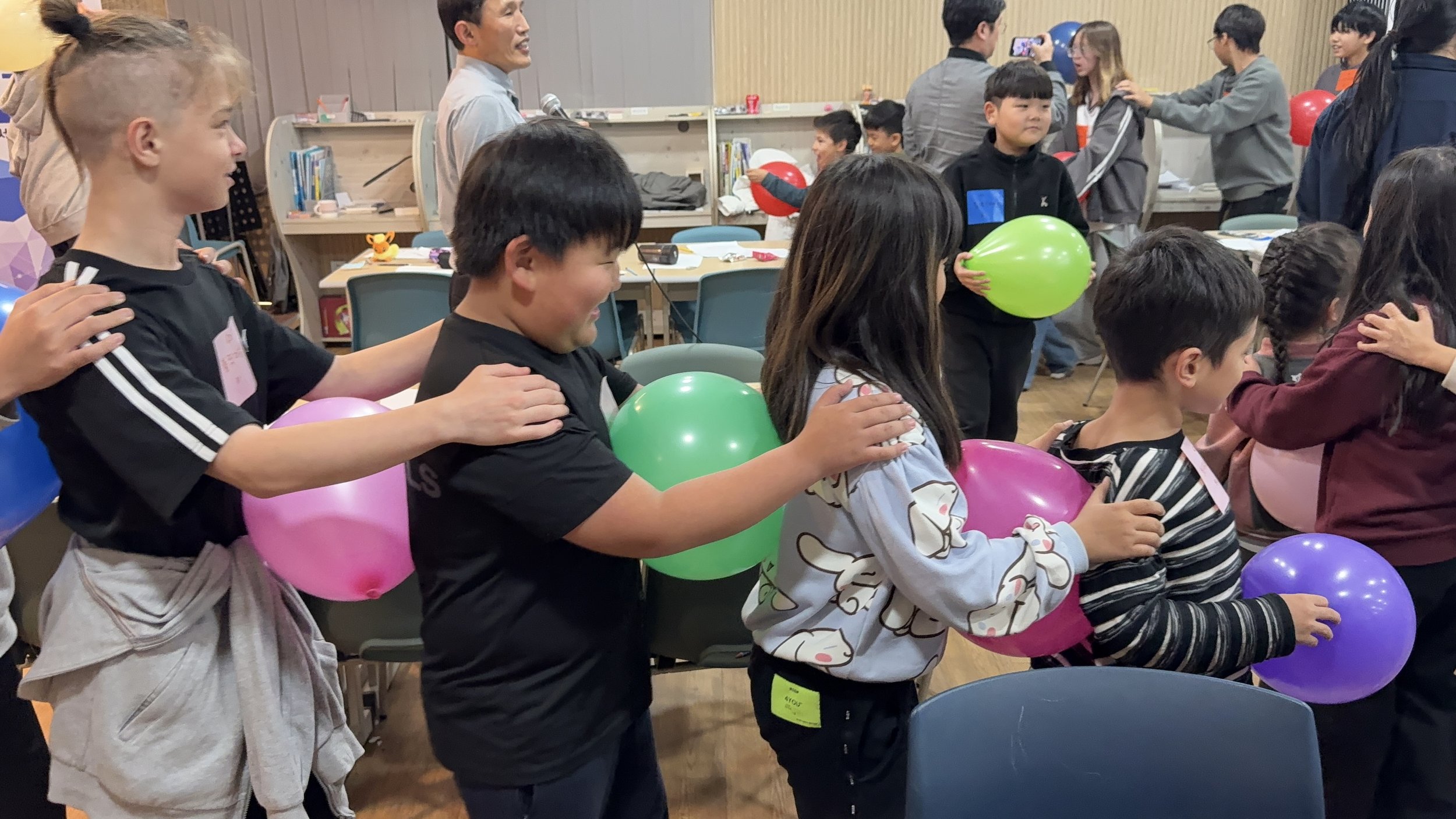 Children participating in a game or activity holding colorful balloons while standing in a line, with several adults in the background.