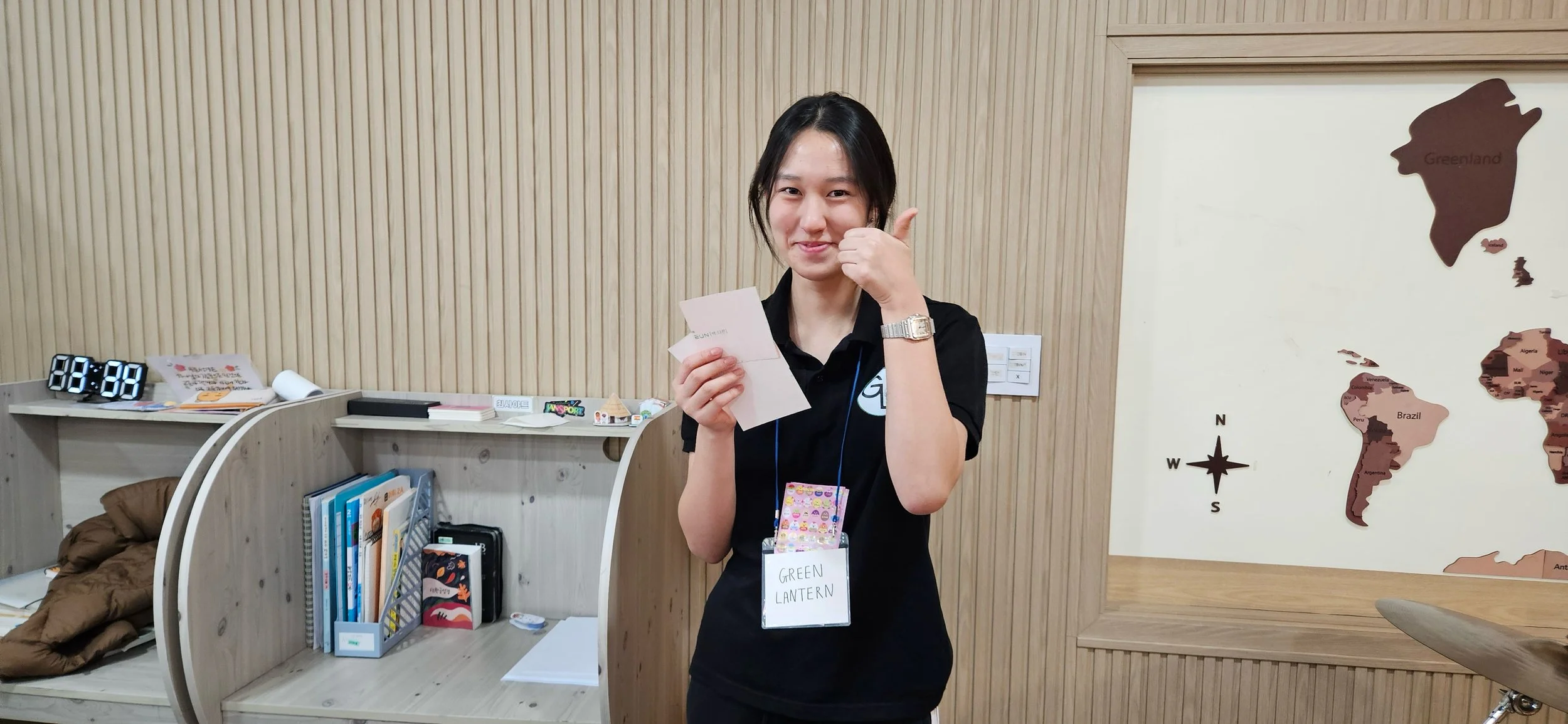 Young woman with updo hairstyle smiling and giving a thumbs up while holding papers, wearing a black polo shirt with a lanyard that says 'GREEN LANTERN' in a room with wooden walls, bookshelves, and a large map of the world.