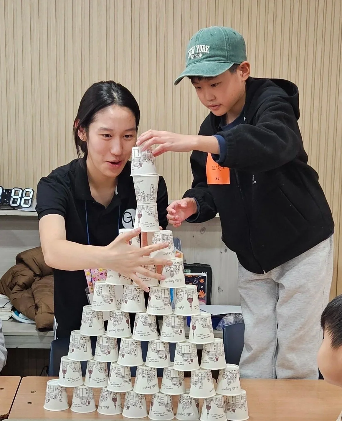 Two children build a pyramid of paper cups with one woman assisting, indoors with a wooden wall background.
