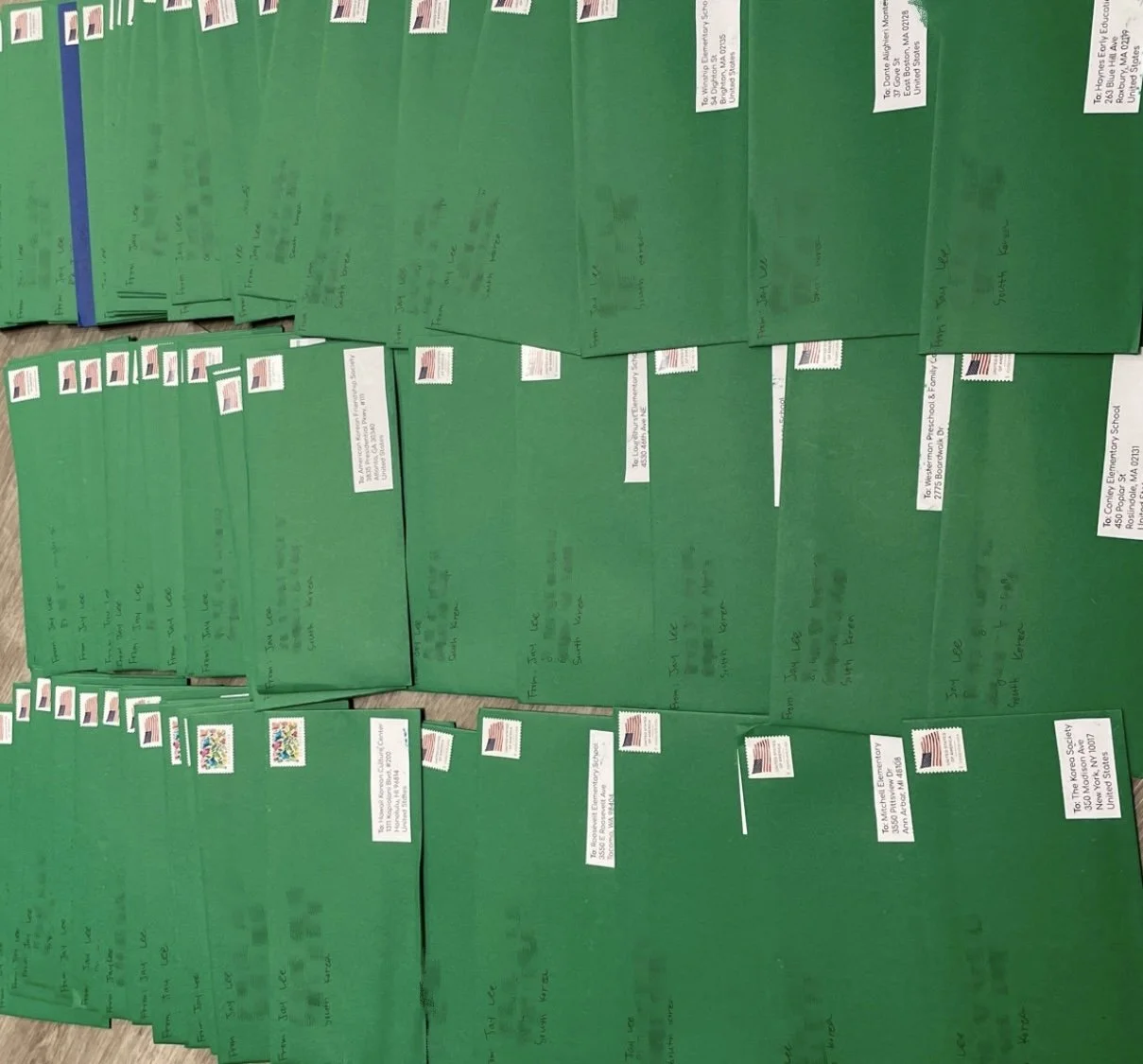 Numerous green envelopes with handwritten addresses and decorated with American flag stamps, arranged on a wooden surface.