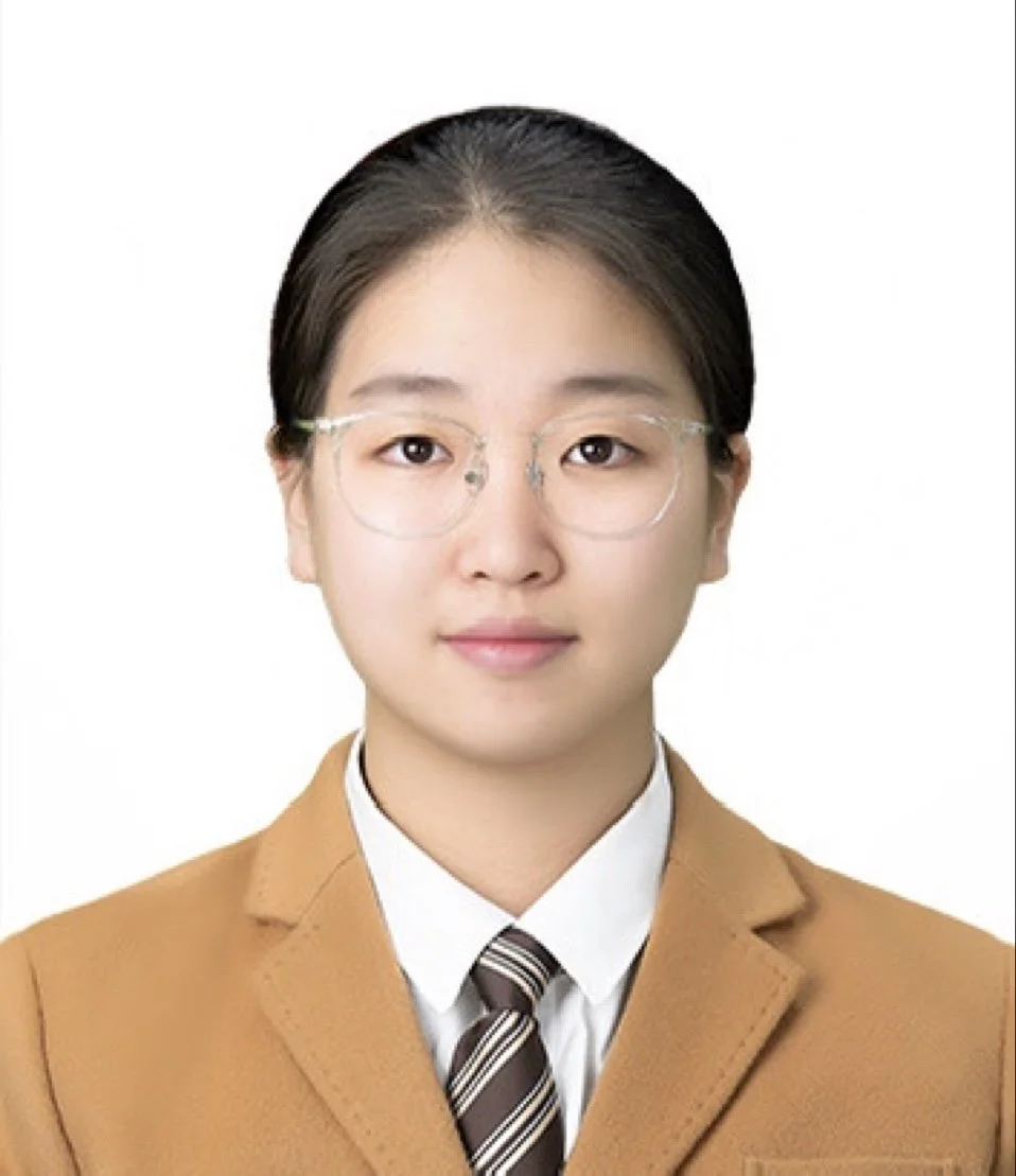 A young woman with glasses, wearing a tan blazer, white shirt, and striped tie, looking directly at the camera against a plain white background.