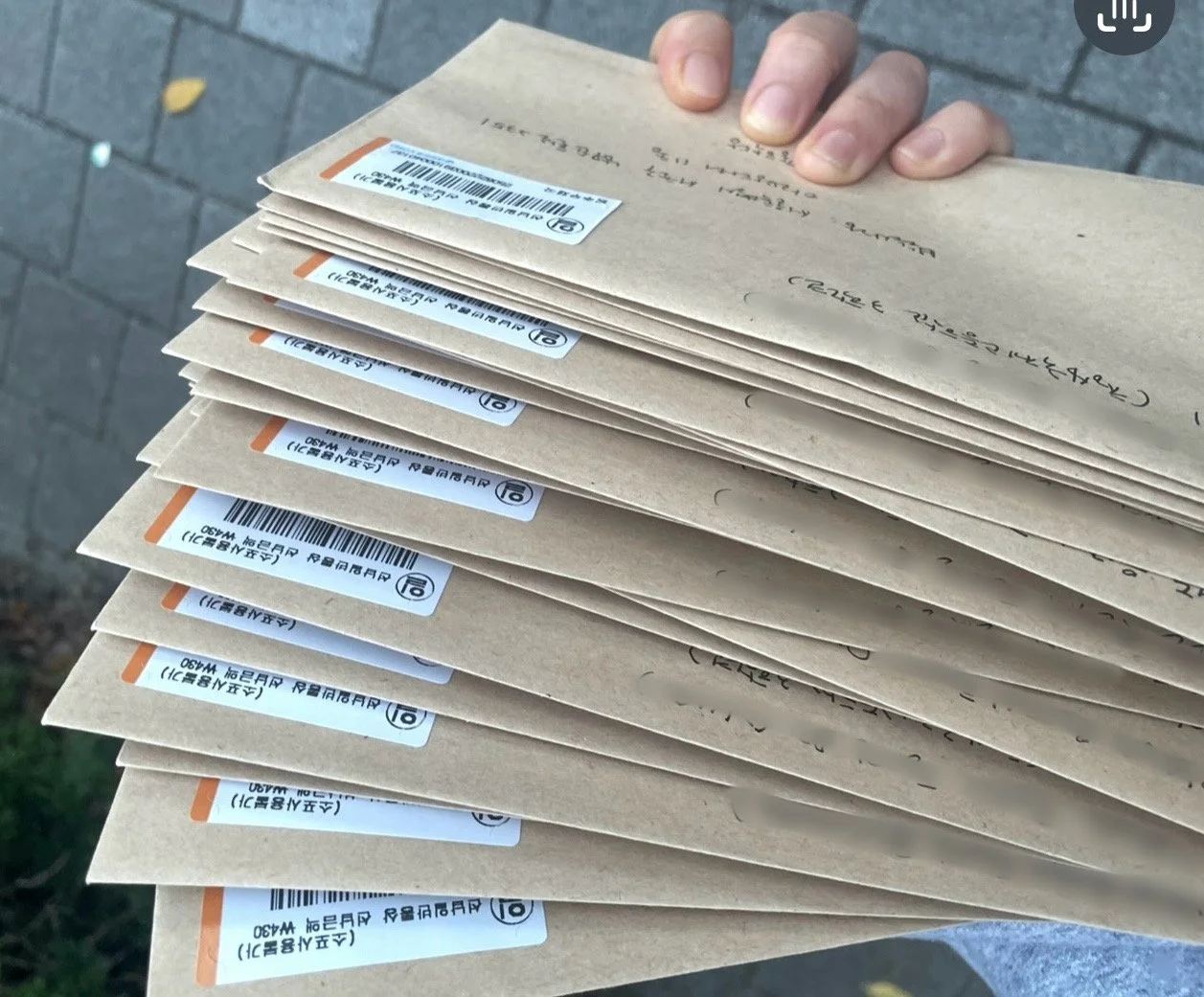 A person holding a stack of brown paper envelopes with barcode labels on each, outdoors on a brick pavement.