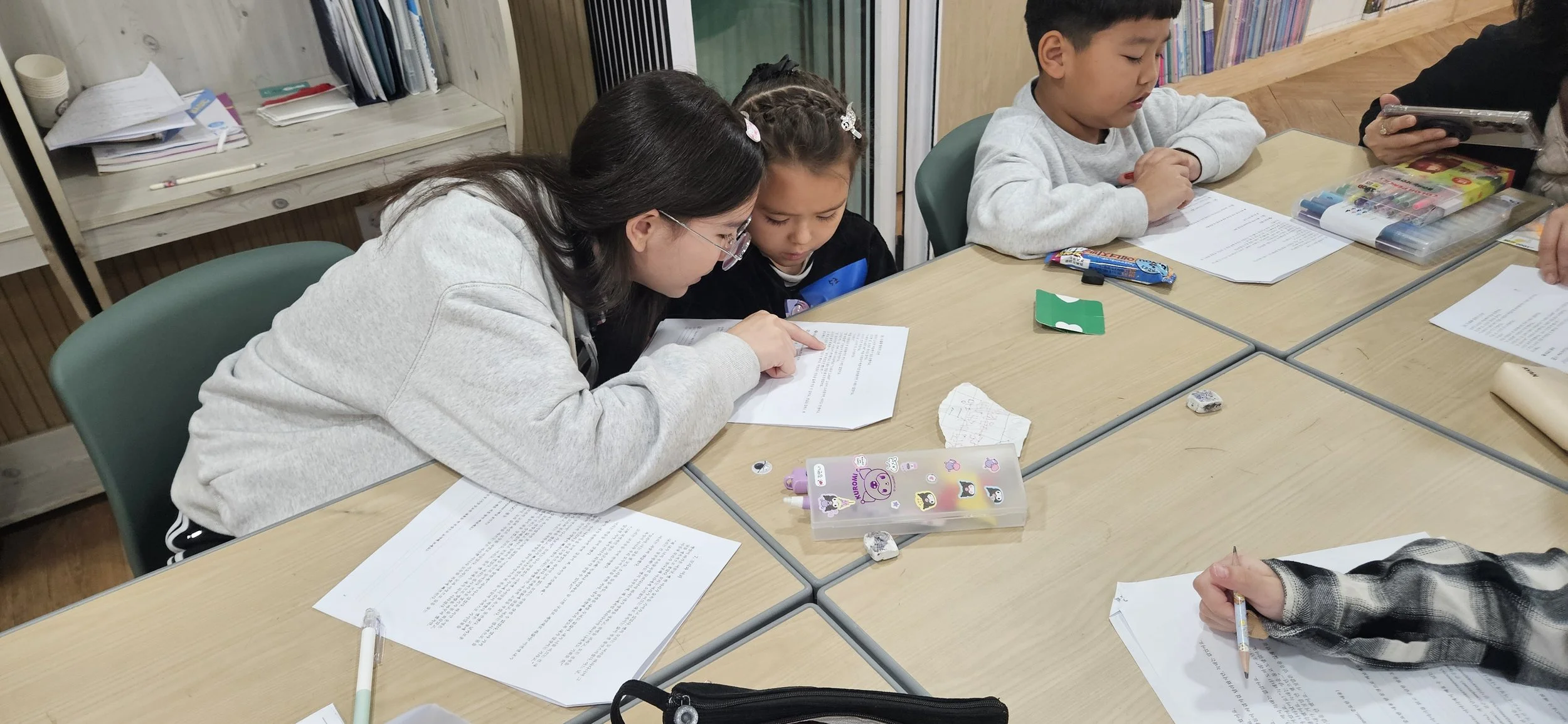 A woman and a young girl sit closely together at a table, looking at a sheet of paper. Other children and papers are on the table, surrounded by school supplies and notebooks.
