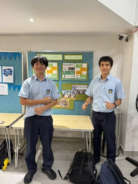Two boys in school uniforms standing in front of a bulletin board in a classroom, smiling, with backpacks on the floor.