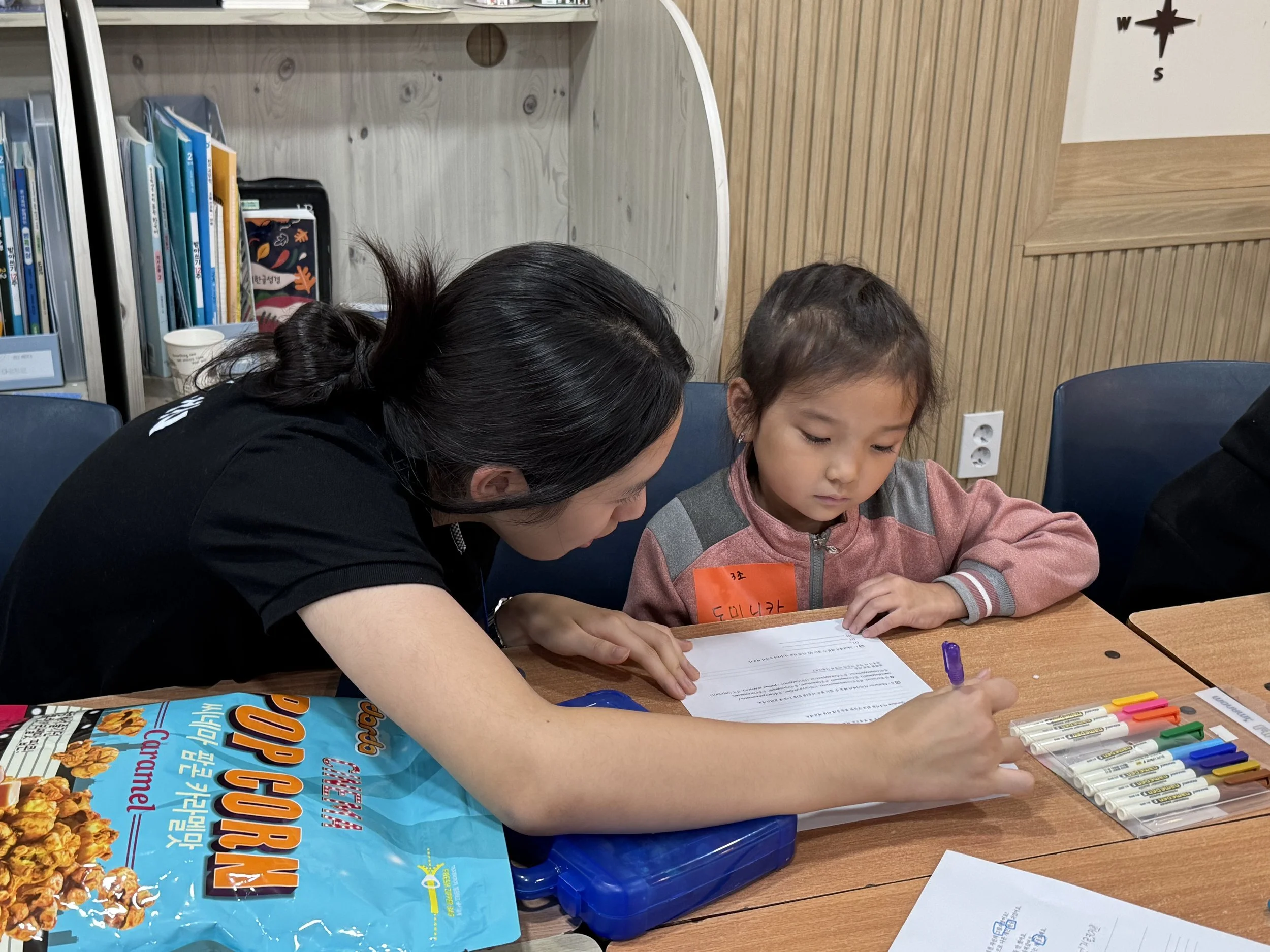 A woman helping a young girl with a worksheet at a table, with school supplies and snacks nearby.