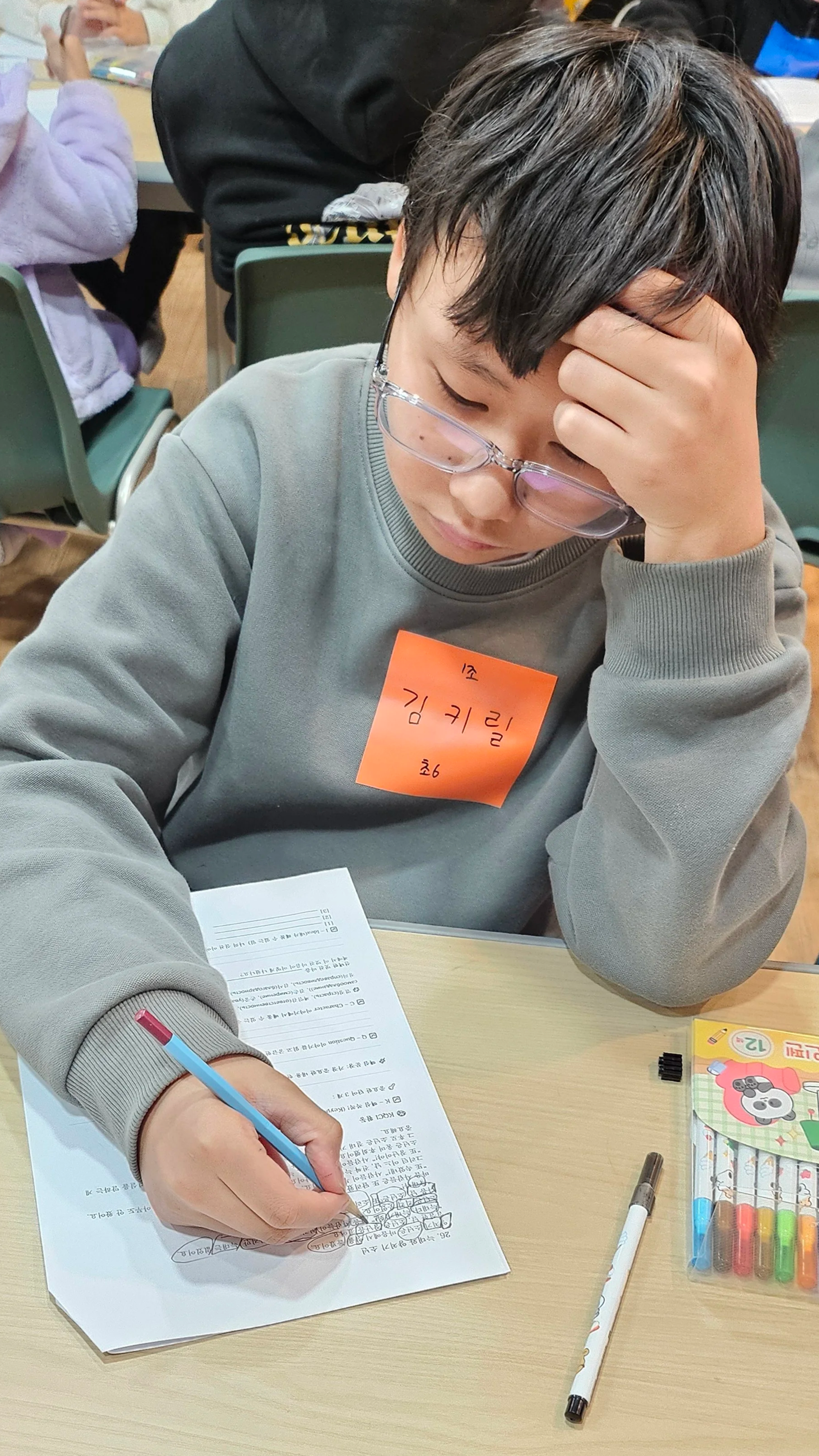 A young boy wearing glasses and a gray sweatshirt, sitting at a desk, writing on a worksheet with a blue pen. He has a bright orange sticky note on his chest with handwritten text in Korean. There are colored markers and a white pen on the desk, and other children are visible in the background, engaged in activities.
