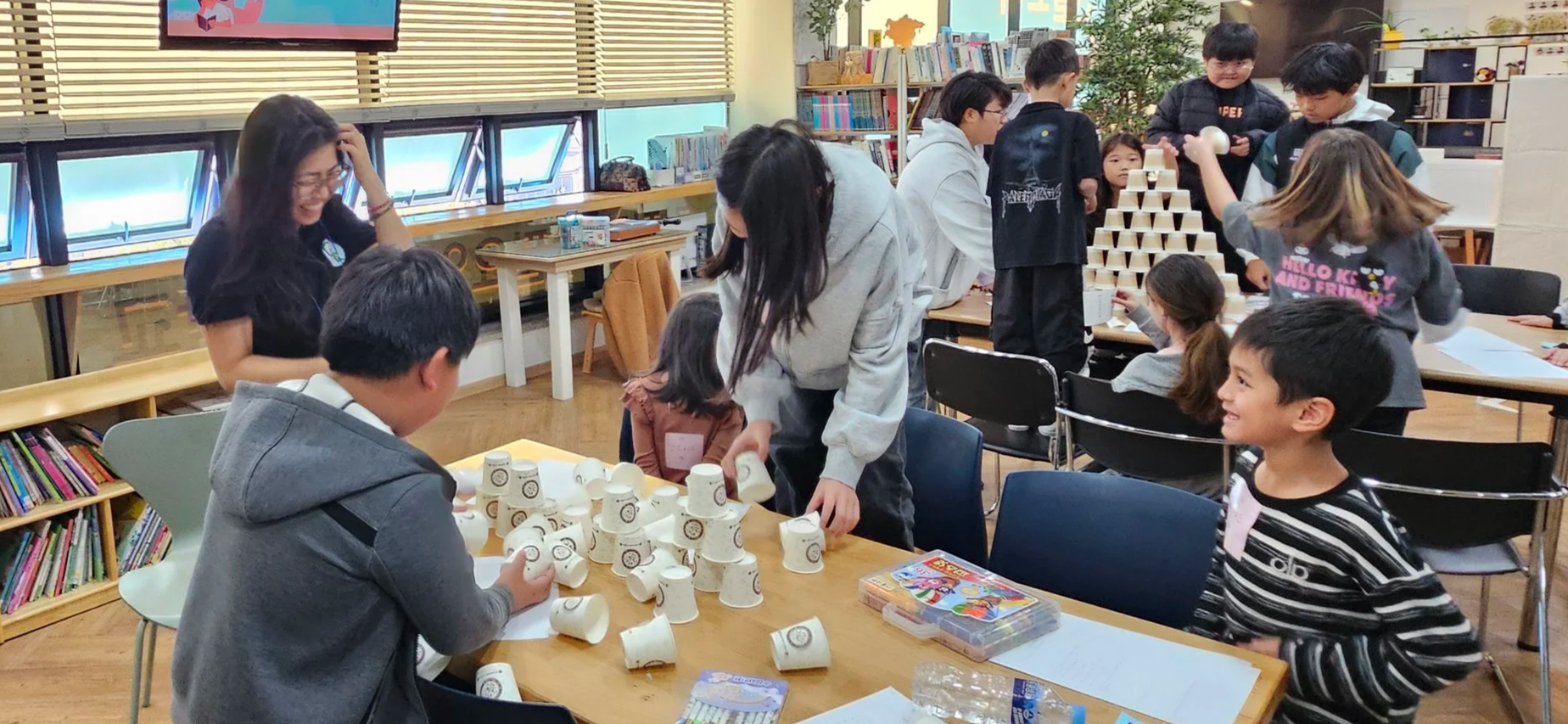 Children and adults playing Jenga with paper cups in a classroom or library setting, with bookshelves and windows in the background.