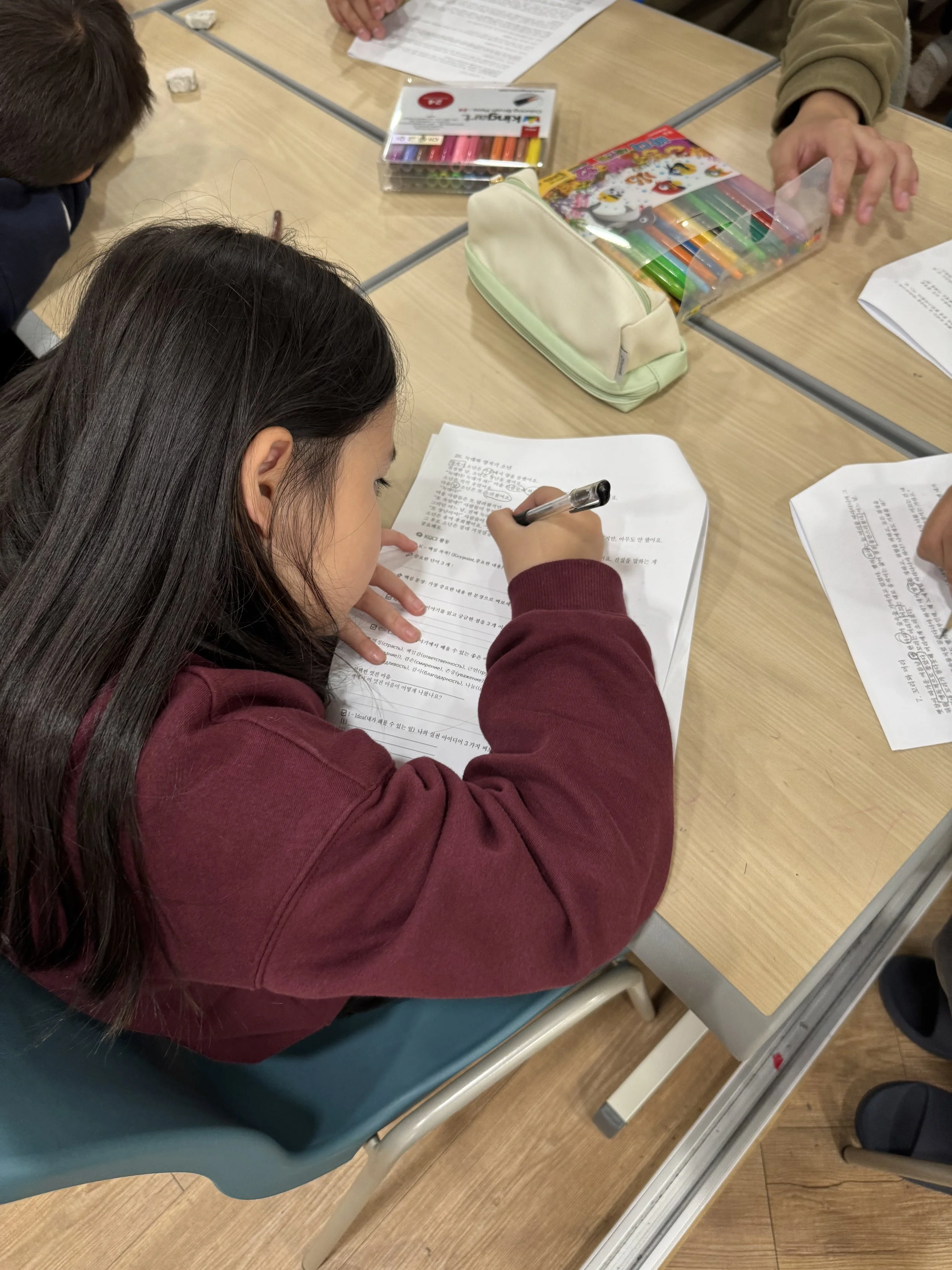 A young girl with long dark hair sitting at a desk, writing on a sheet of paper with printed text. There are school supplies, including a colorful box of pens or markers, a white pencil case, and some papers on the desk.