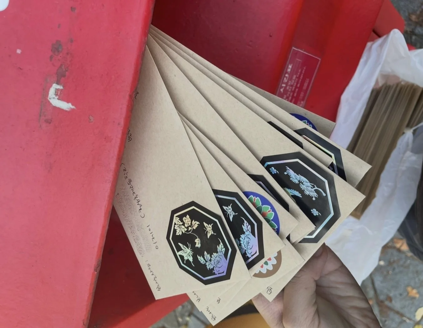 Person holding several paper bags, some with colorful designs, next to a red surface and a white plastic bag filled with more paper bags in the background.