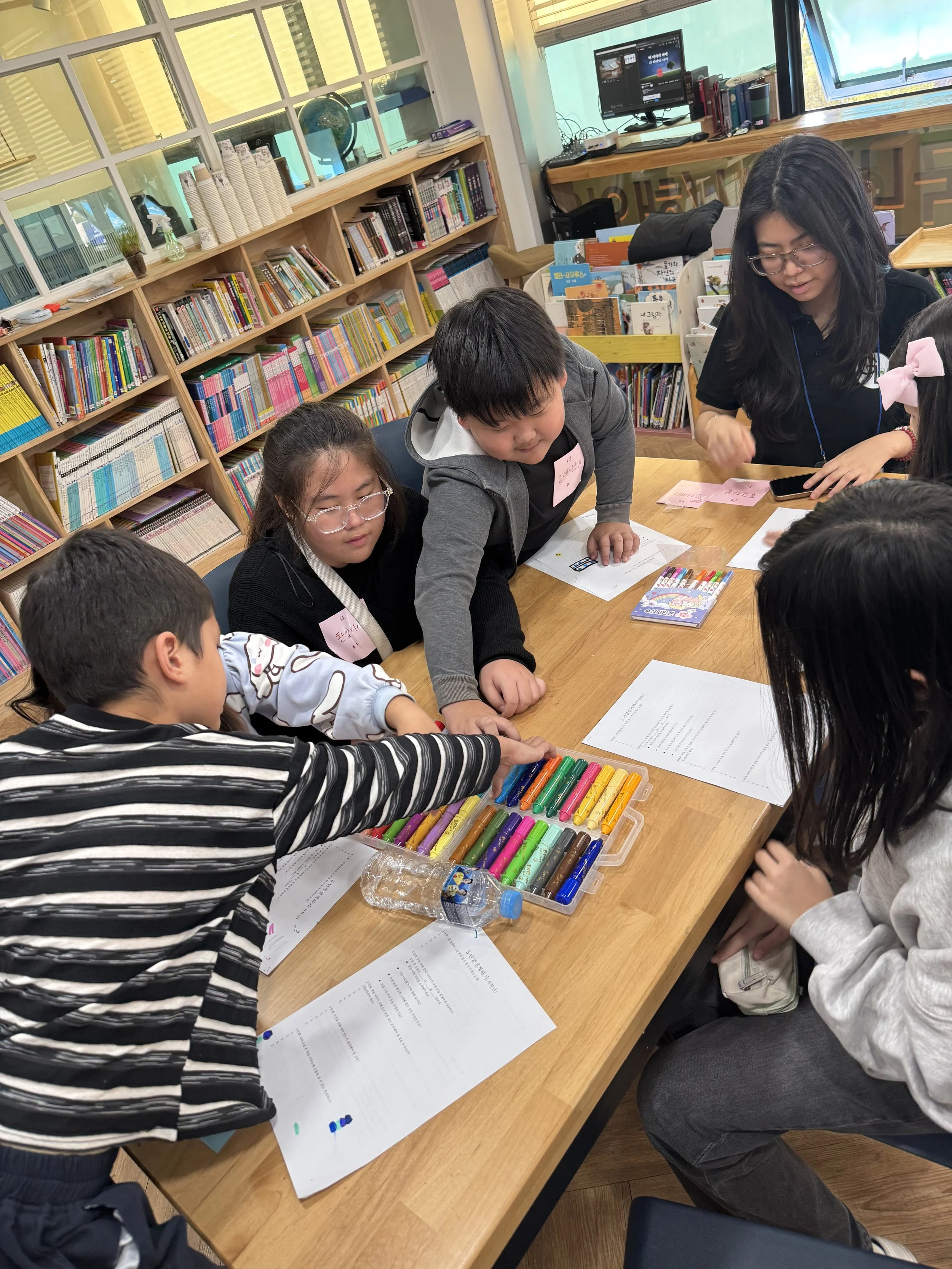 Children and a woman gathered around a table with colored chalks, papers, and a water bottle in a library or classroom environment.
