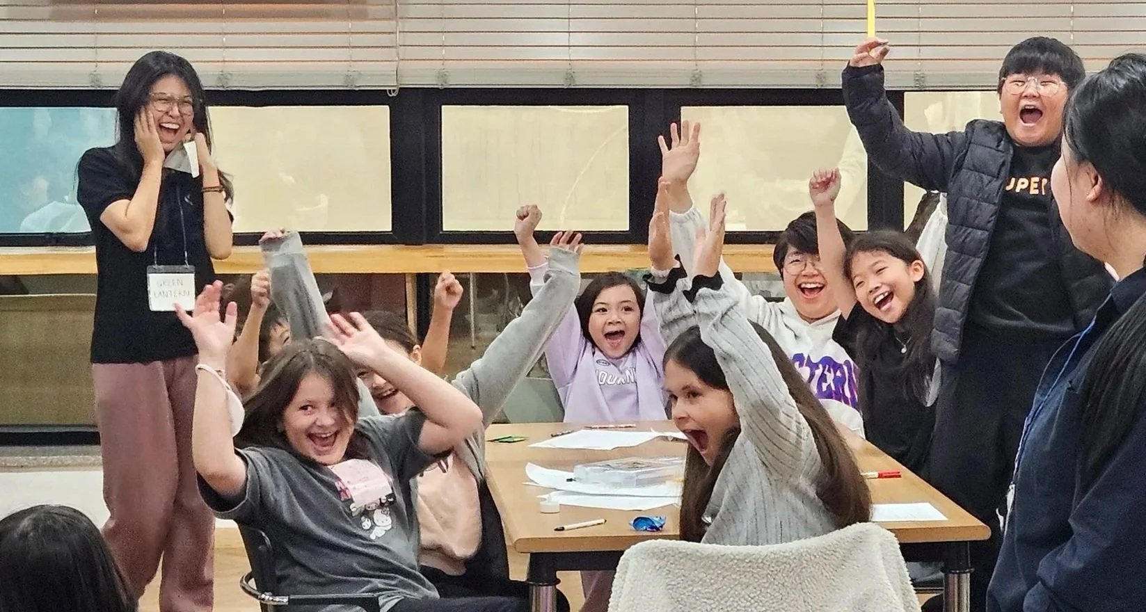 Children and adults in a classroom excitedly celebrating or playing a game, some with raised hands and wide smiles.