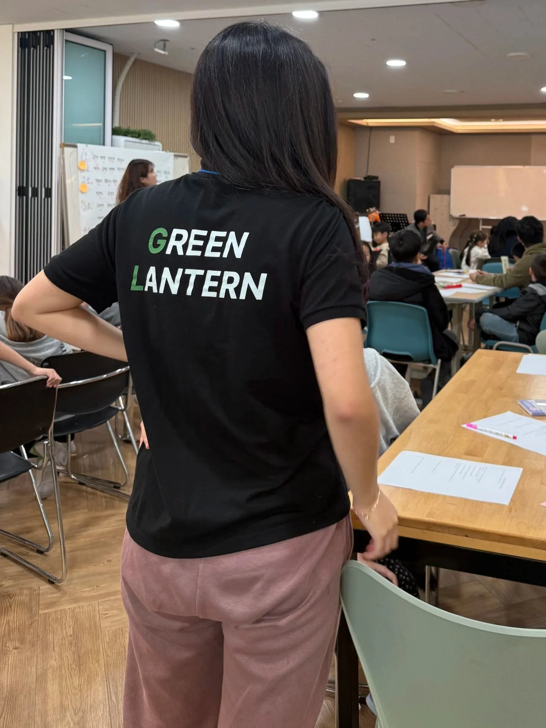 A woman wearing a black T-shirt with the words 'GREEN LANTERN' printed on the back, standing in a classroom or conference room filled with students seated at tables.