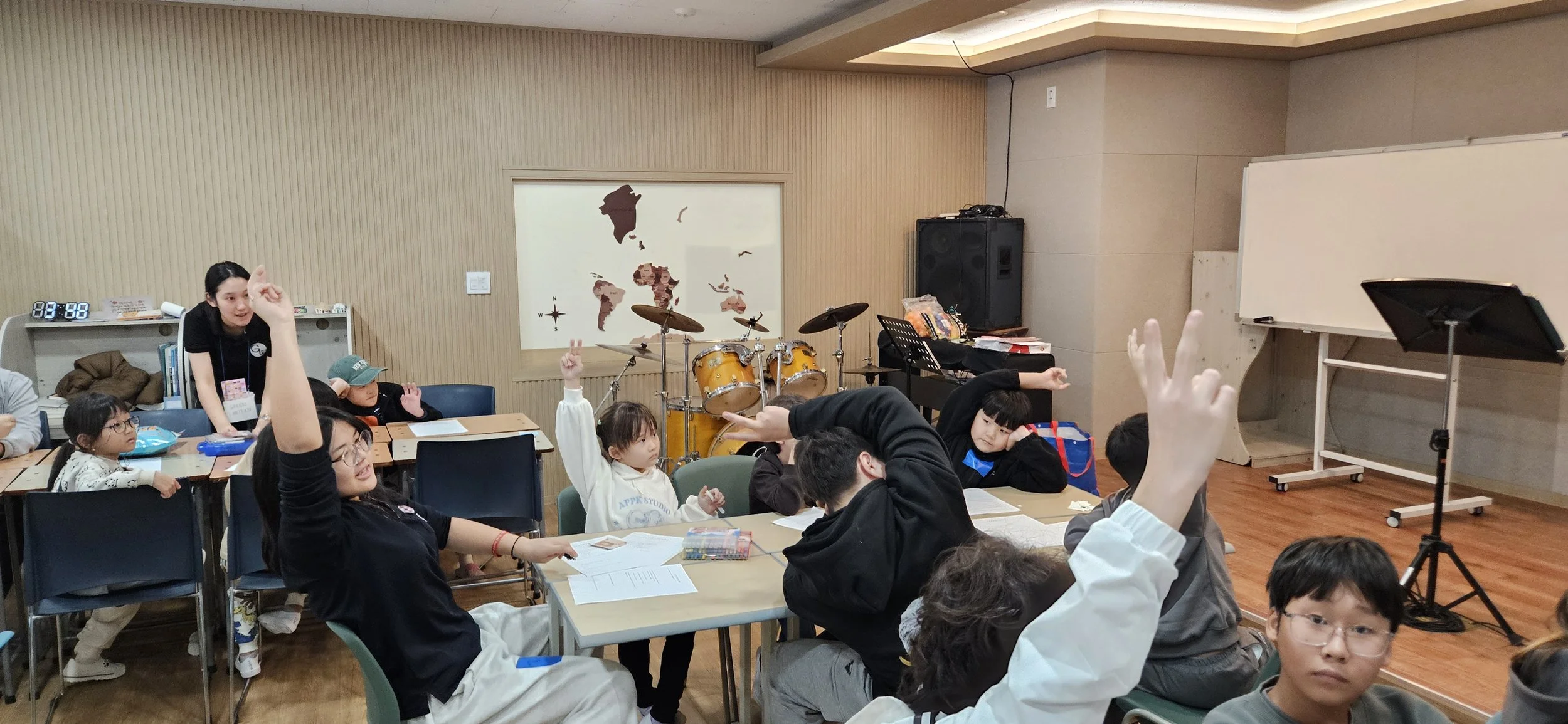 Classroom with children and adults participating in a discussion or activity, some raising their hands, in a music room with drums and a whiteboard.