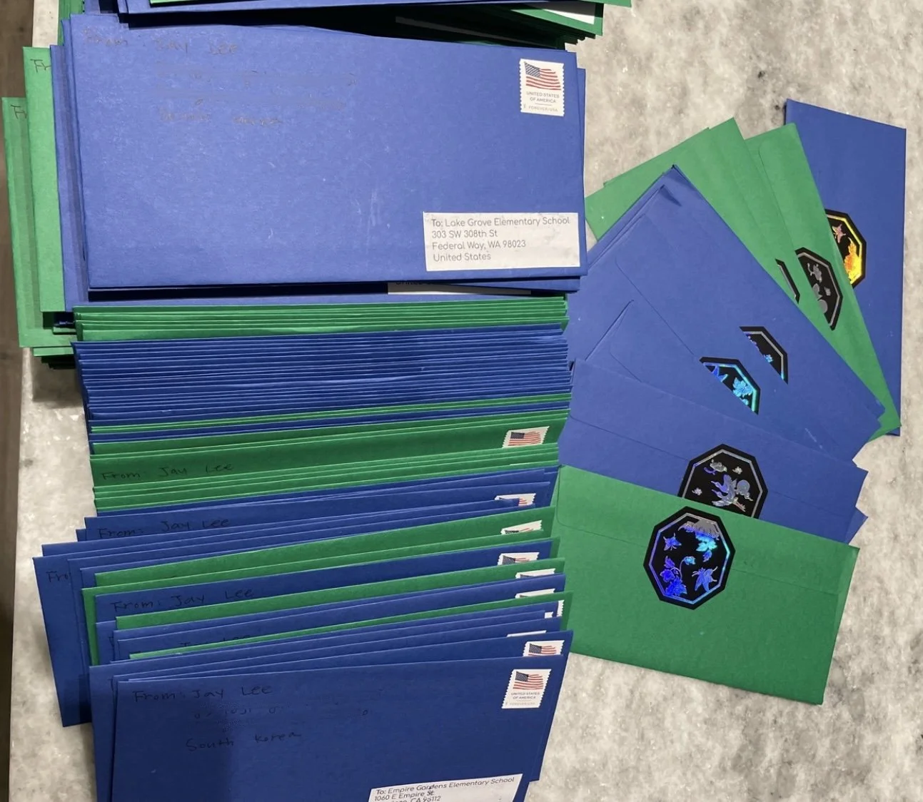 Stacks of blue and green envelopes with stickers and handwritten addresses, arranged on a gray surface, some envelopes showing holographic and printed stamps.