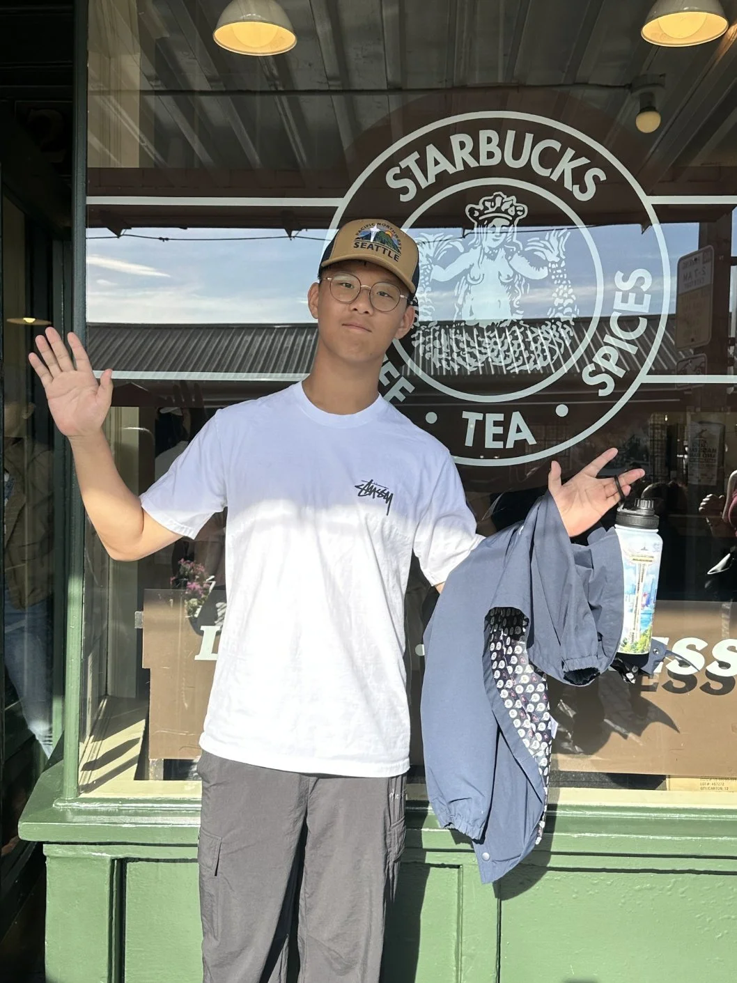 Young man standing outside Starbucks coffee shop, waving with his right hand, holding a jacket in his left hand, wearing glasses, a beige cap with 'SEATTLE' logo, and a white t-shirt.
