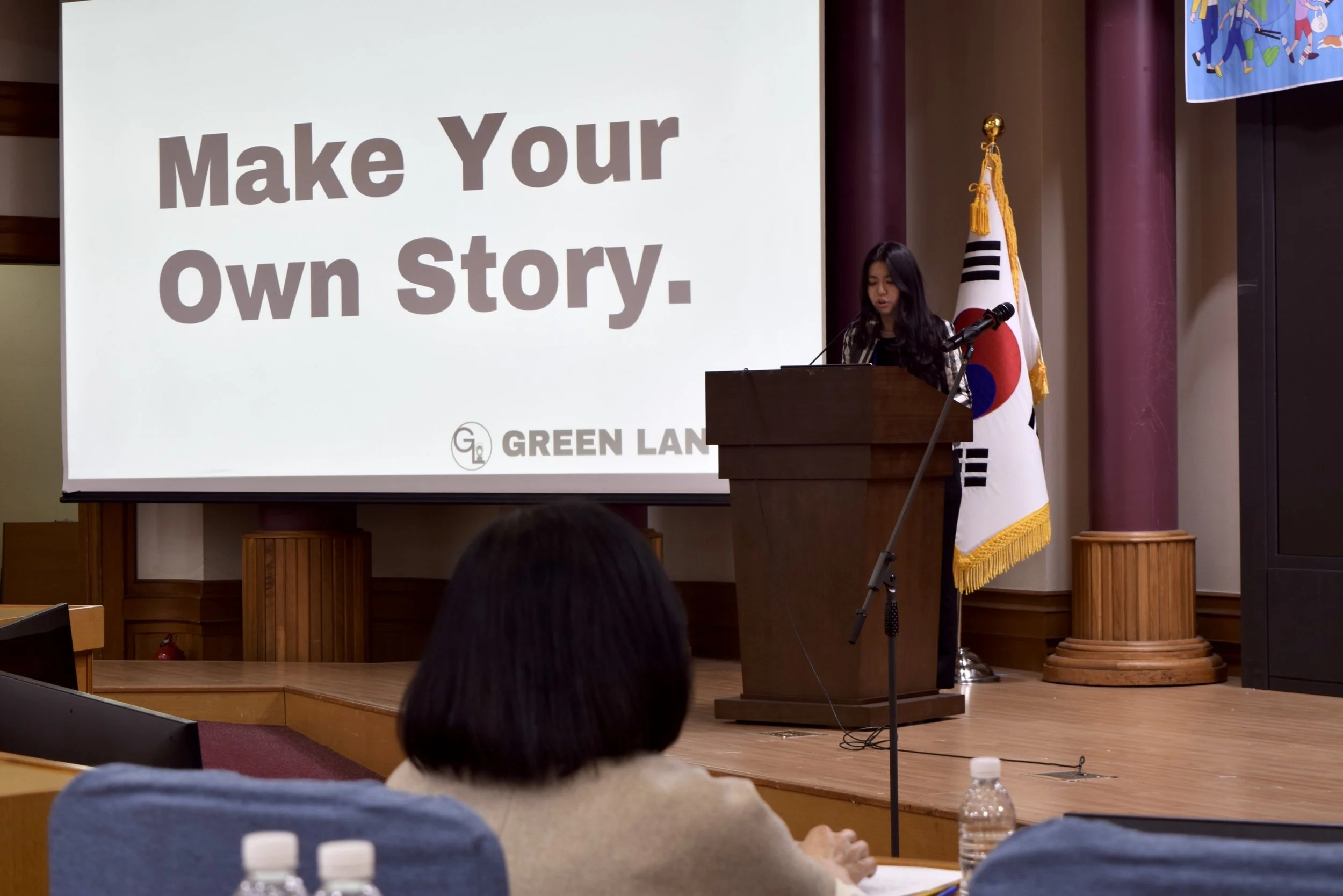 A young woman giving a presentation at a podium in a conference room, with a large screen behind her displaying the words 'Make Your Own Story.' and a logo reading 'GREEN LAN.' There is a South Korean flag behind her, and an audience member is visible in the foreground.
