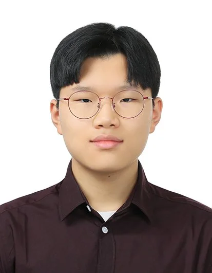 A young person with straight black hair, wearing glasses and a dark button-up shirt, looking directly at the camera against a plain white background.