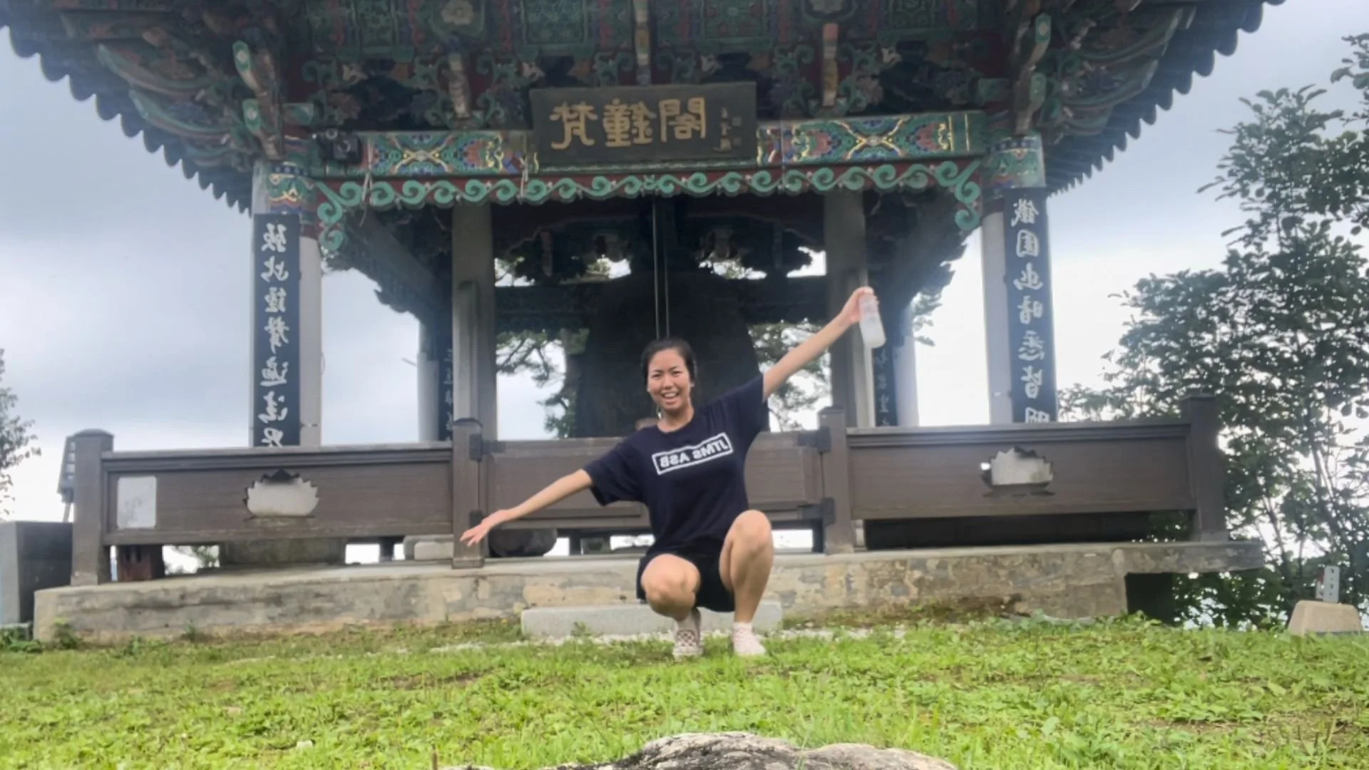 A smiling woman crouches down with arms outstretched in front of a traditional Asian pavilion with intricate wood carvings and Chinese characters on signs, surrounded by greenery and cloudy sky.