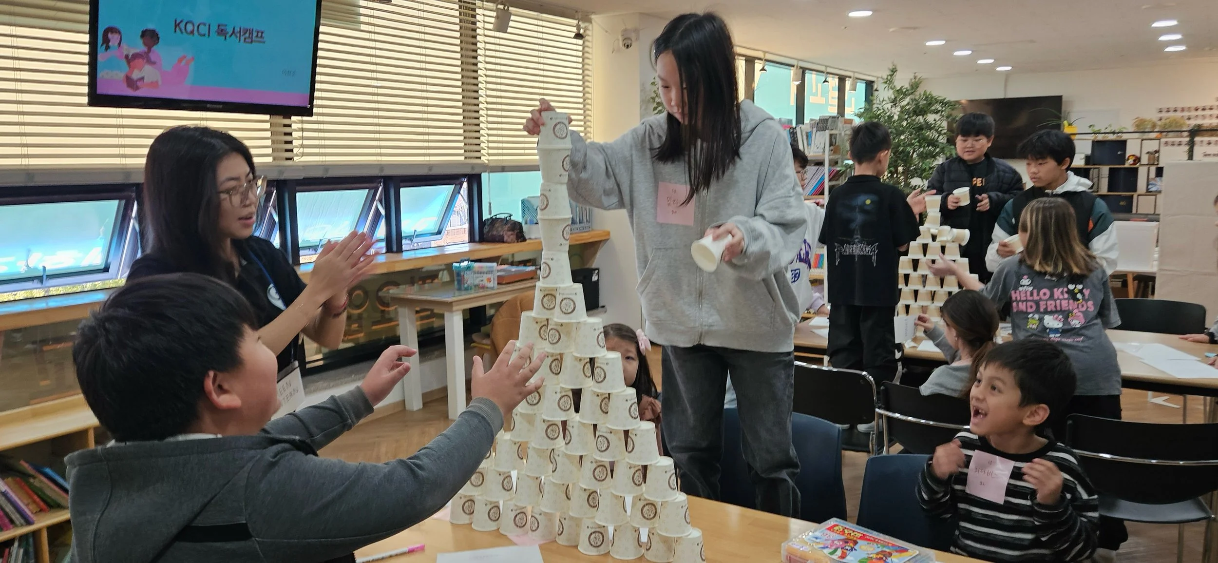 Children building a pyramid with paper cups in a classroom or library setting, with some children smiling and engaging in the activity.