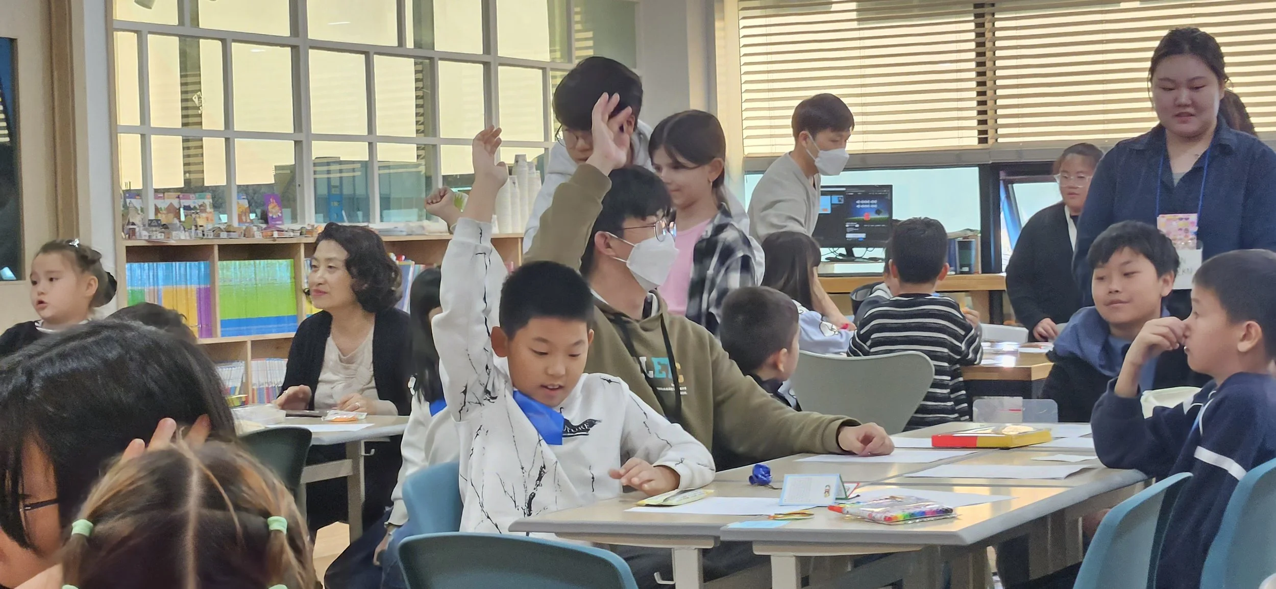 Children and adults in a classroom, some children raising their hands, some seated at tables with books and supplies, with bookshelves and computer monitors in the background.